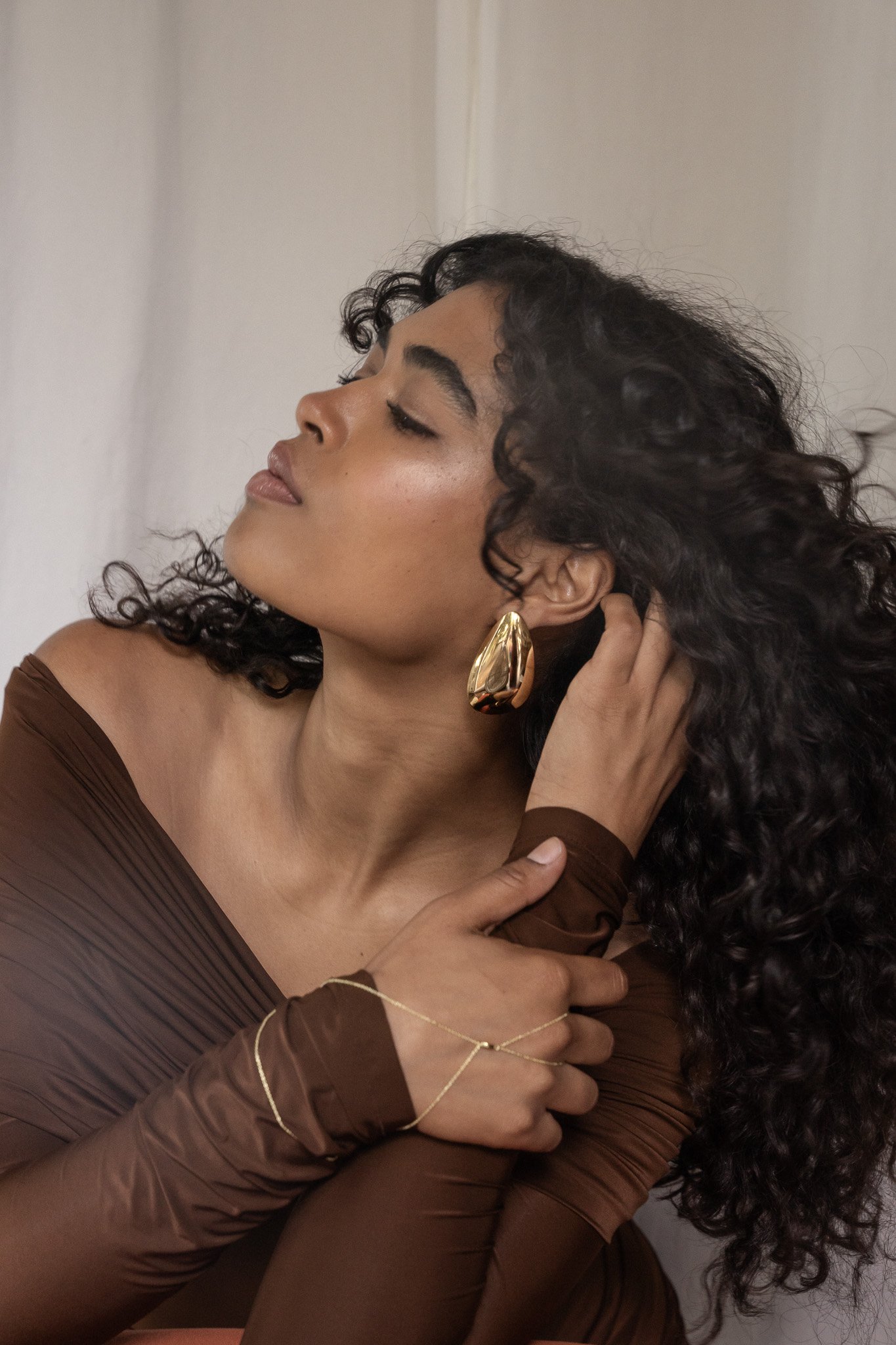 Woman with curly hair wearing large gold earring and brown off-shoulder top.