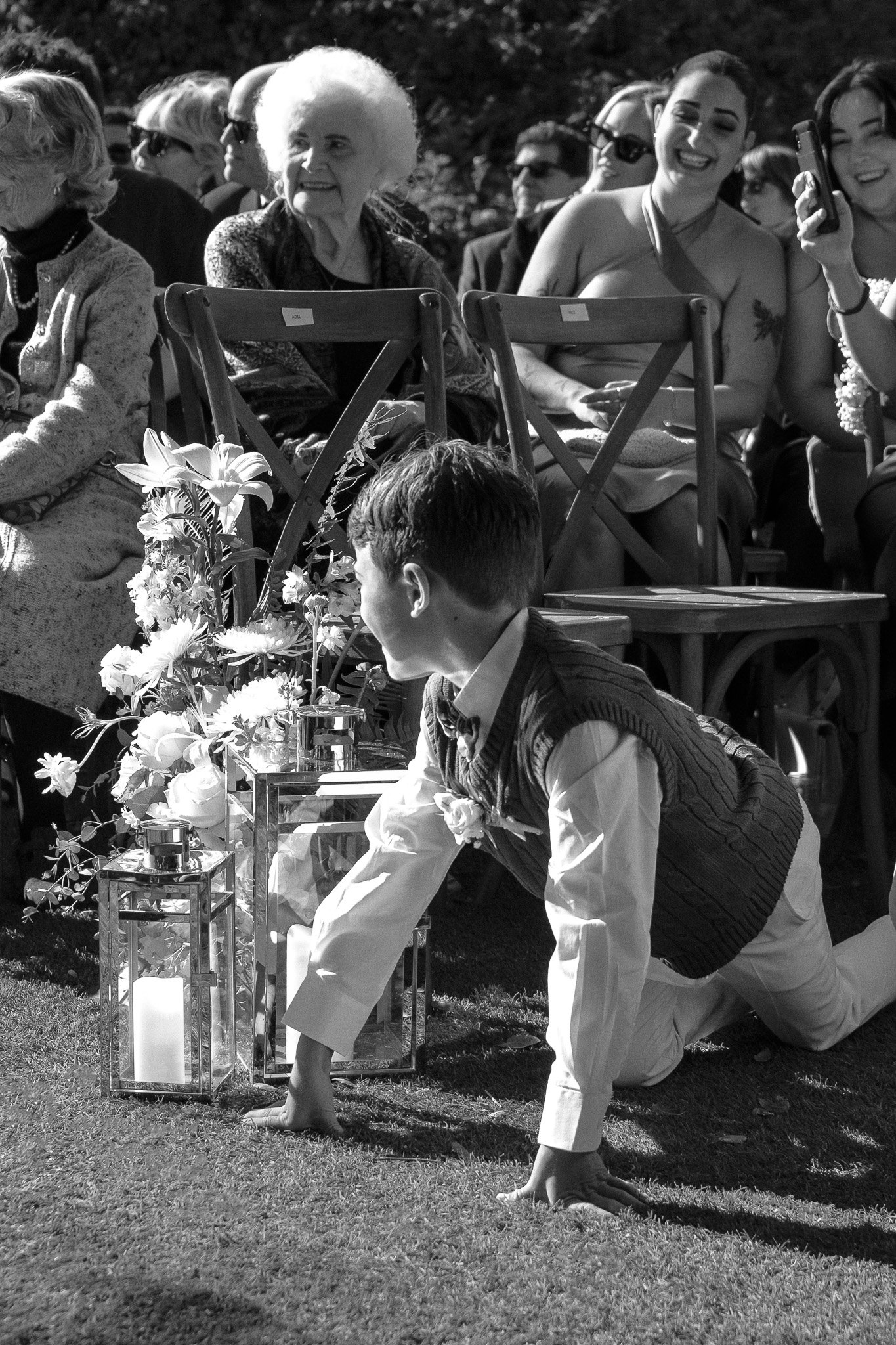 A young boy in formal attire is kneeling on the grass near decorative lanterns and flowers at an outdoor event. He is surrounded by seated adults, some of whom are smiling and taking photos. The scene appears to be from a wedding or similar celebrati