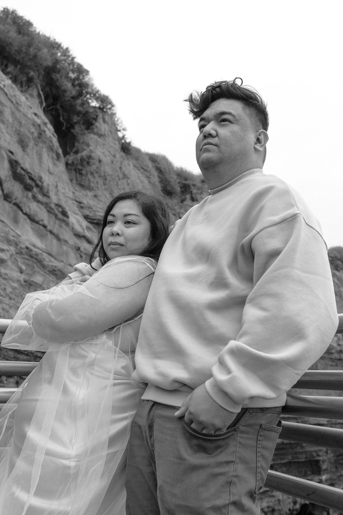 A black and white photo of a couple standing closely together. The woman is wearing a sheer dress, and the man is wearing a sweatshirt. They are outdoors near rocky cliffs. The expression on their faces is calm and contemplative.