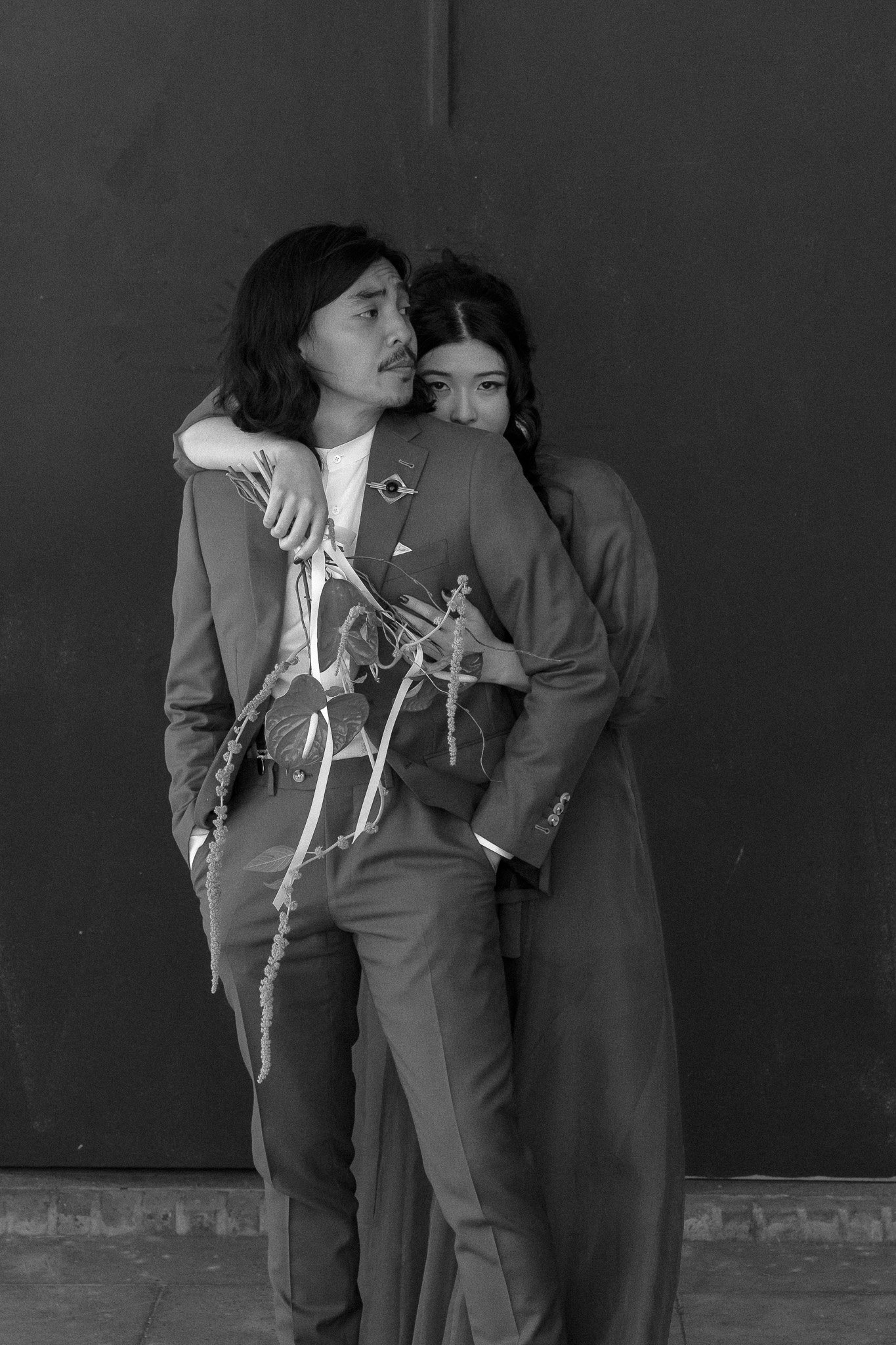 Black and white photo of two people in formal attire, with one embracing the other from behind while holding a bouquet.