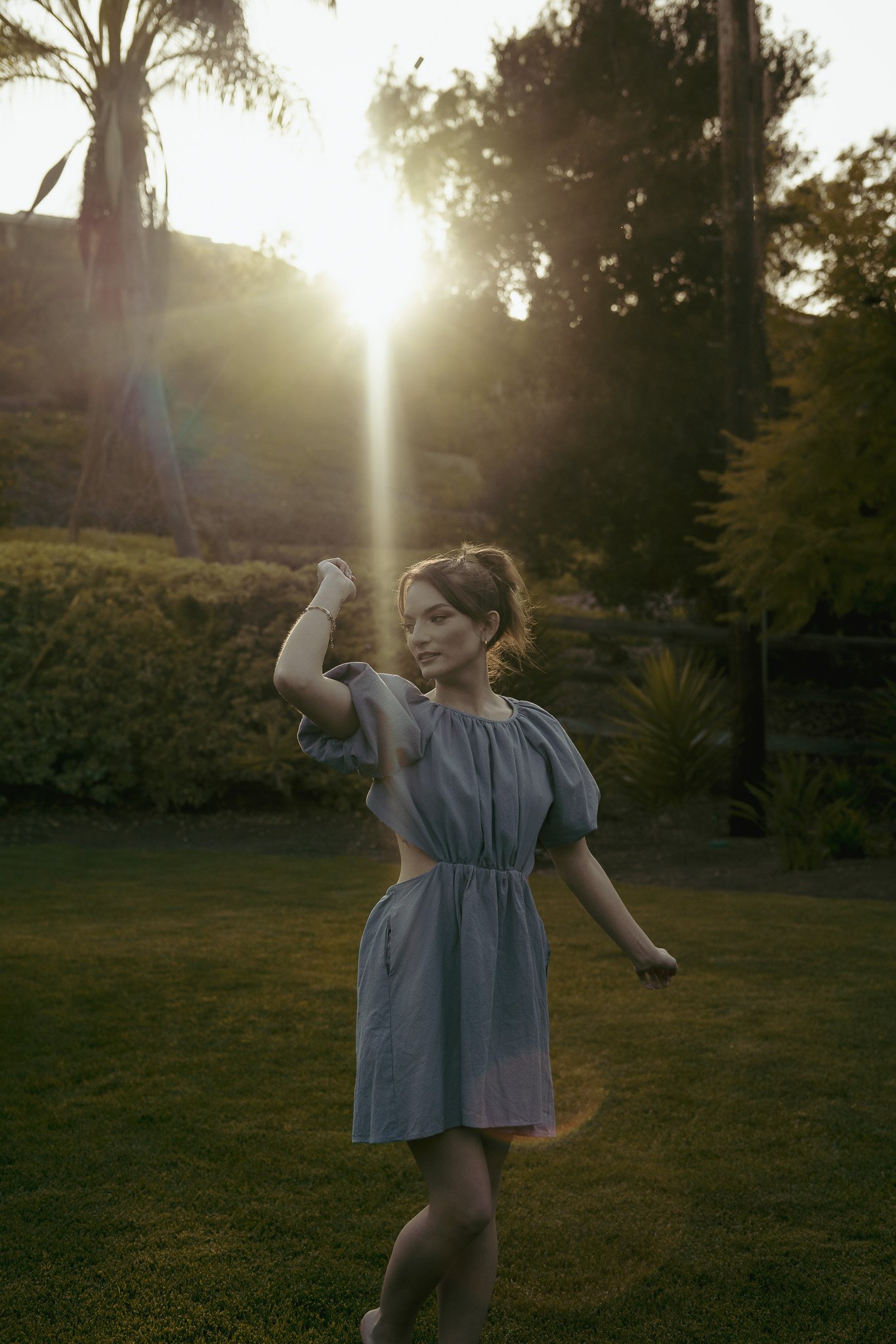 Woman in a light dress dancing on grass during sunset with trees in the background.