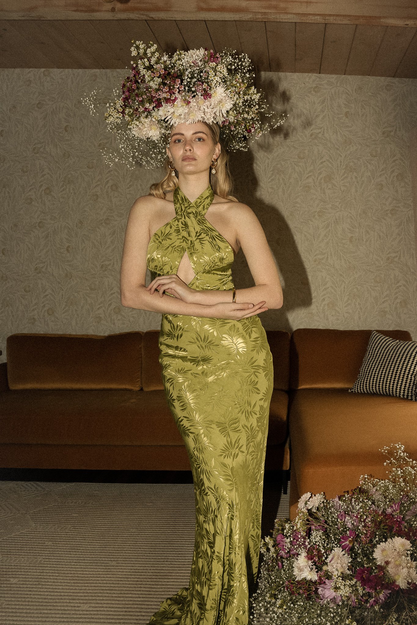 A woman in a green patterned dress stands indoors wearing a large floral headdress, with a bouquet of flowers nearby and a brown sofa in the background.