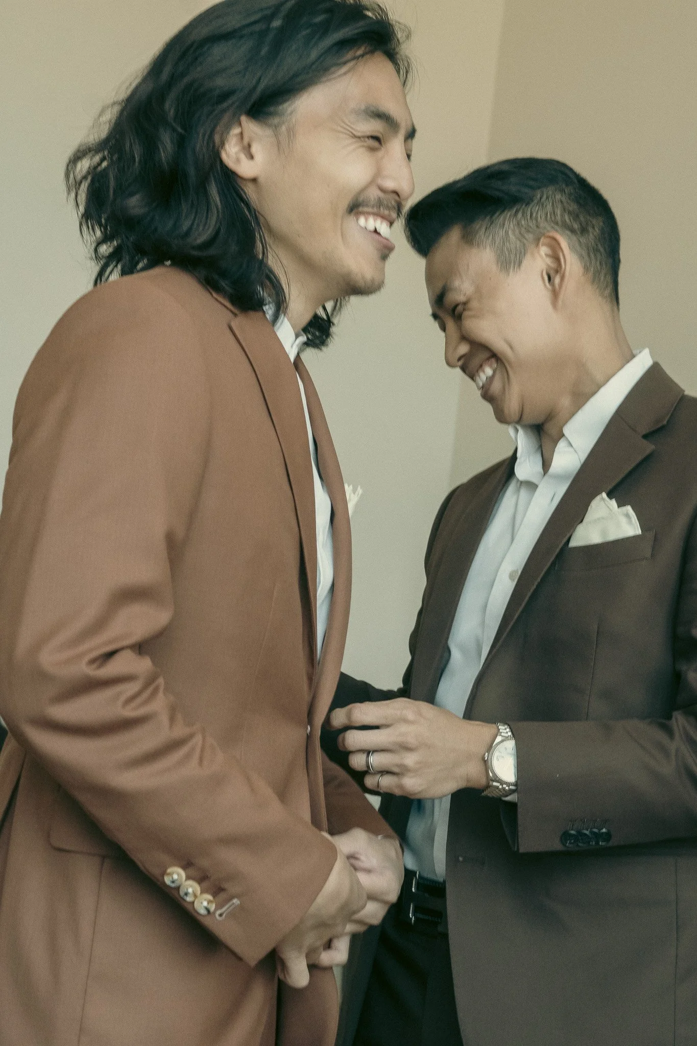 Two men wearing brown suits smiling and interacting, one adjusting the other's jacket, both appear happy.