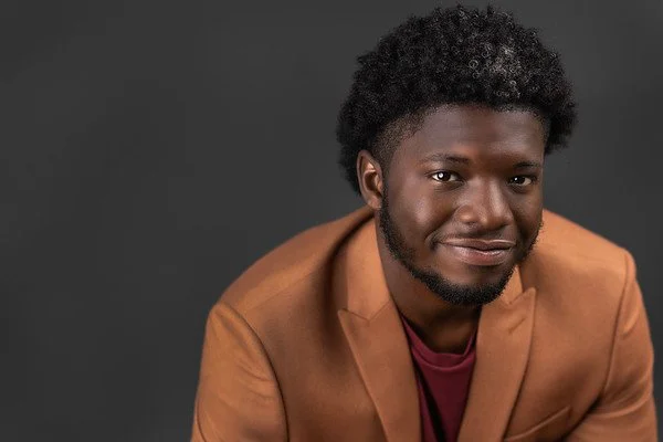 Portrait of a young man with curly hair and a beard, wearing a brown blazer and maroon shirt, smiling against a dark grey background.