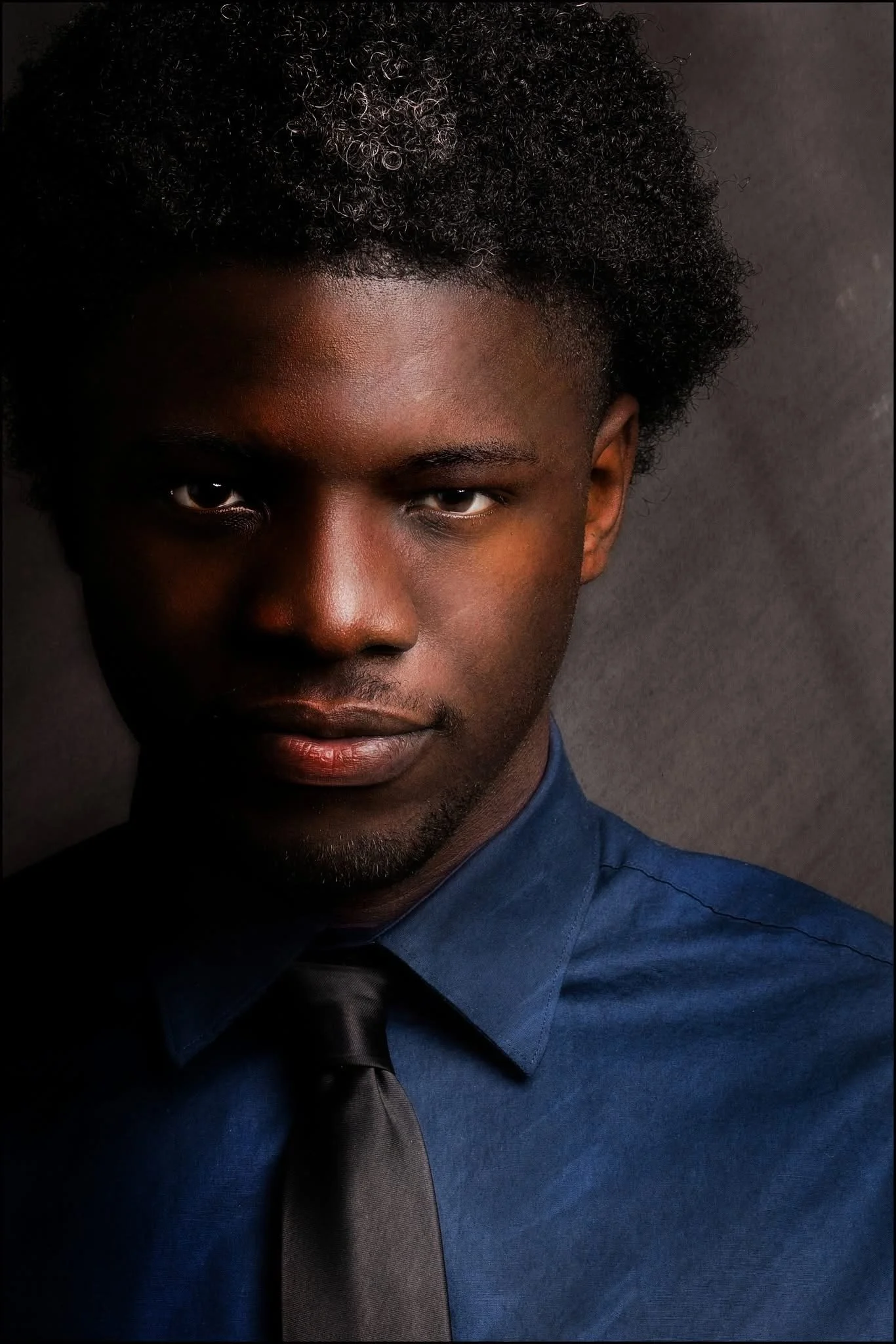 Portrait of a young Black man in a blue shirt and black tie with serious expression, dark background.