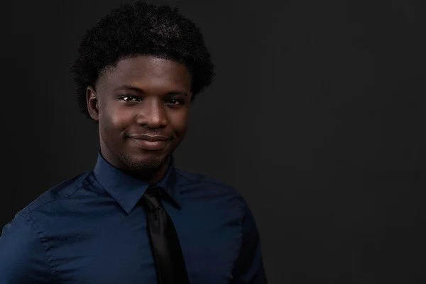Portrait of a young man with curly hair, wearing a dark blue shirt and black tie, against a black background.