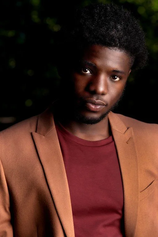 Portrait of a young man with dark curly hair, wearing a brown blazer over a maroon shirt, against a dark outdoor background.