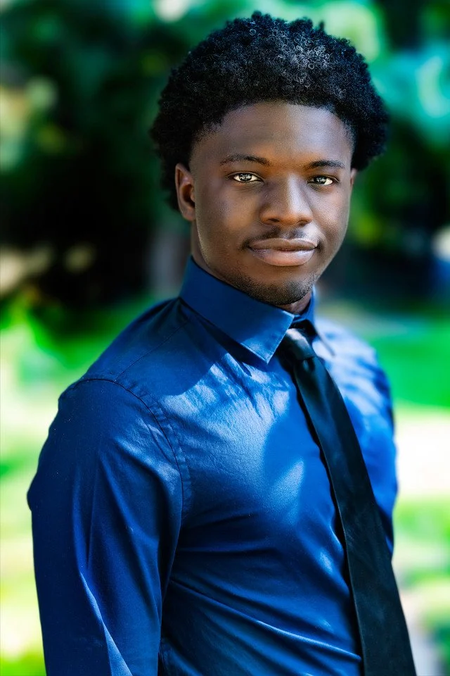 Young man with curly hair wearing a shiny blue dress shirt and a dark tie outdoors with a blurred green background.