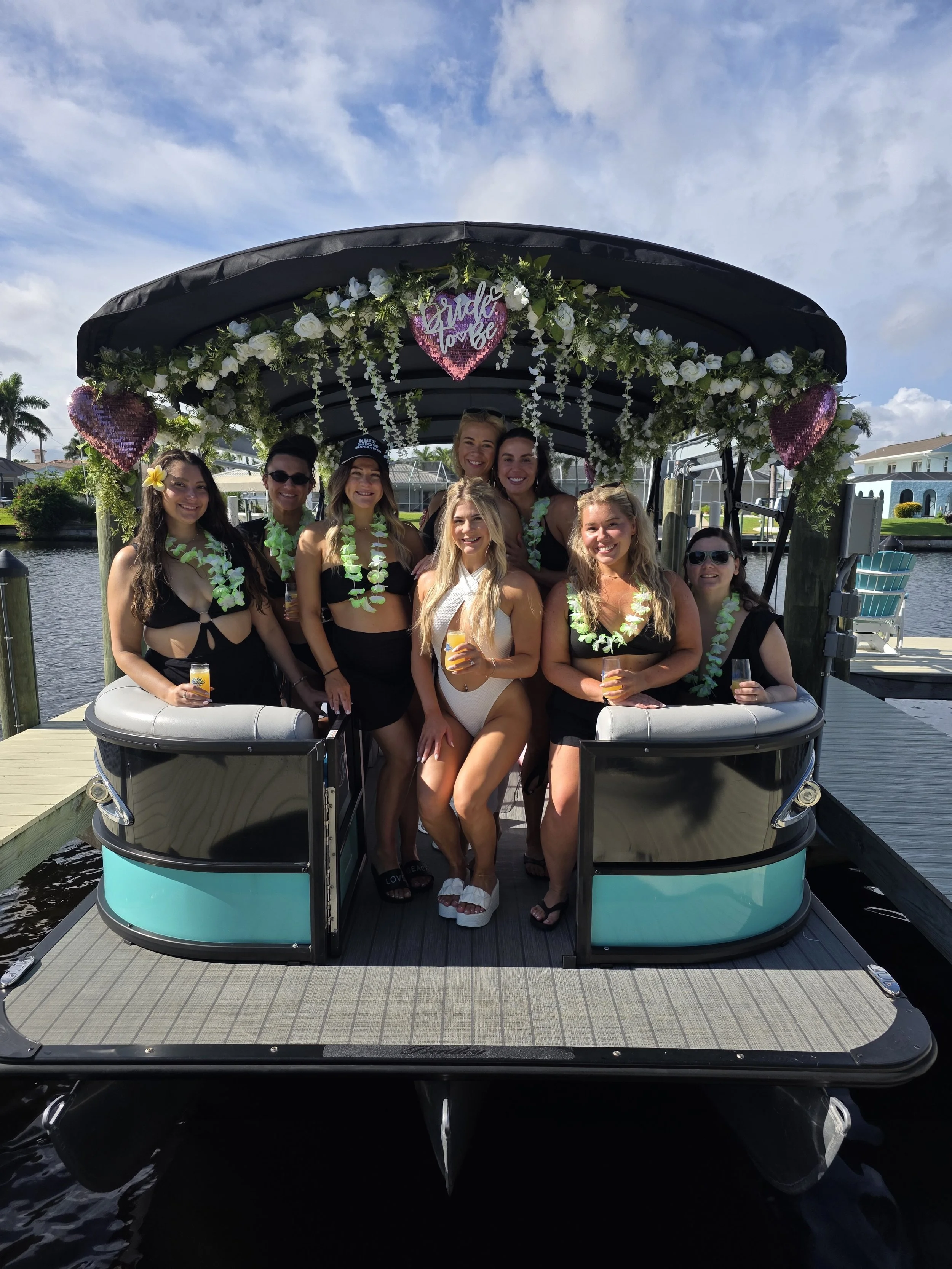 A group of women celebrating a bachelorette party on a boat decorated with white flowers, green foliage, and pink heart-shaped signs that say 'Bride to be.' They are smiling and holding drinks, with a waterway and houses in the background.