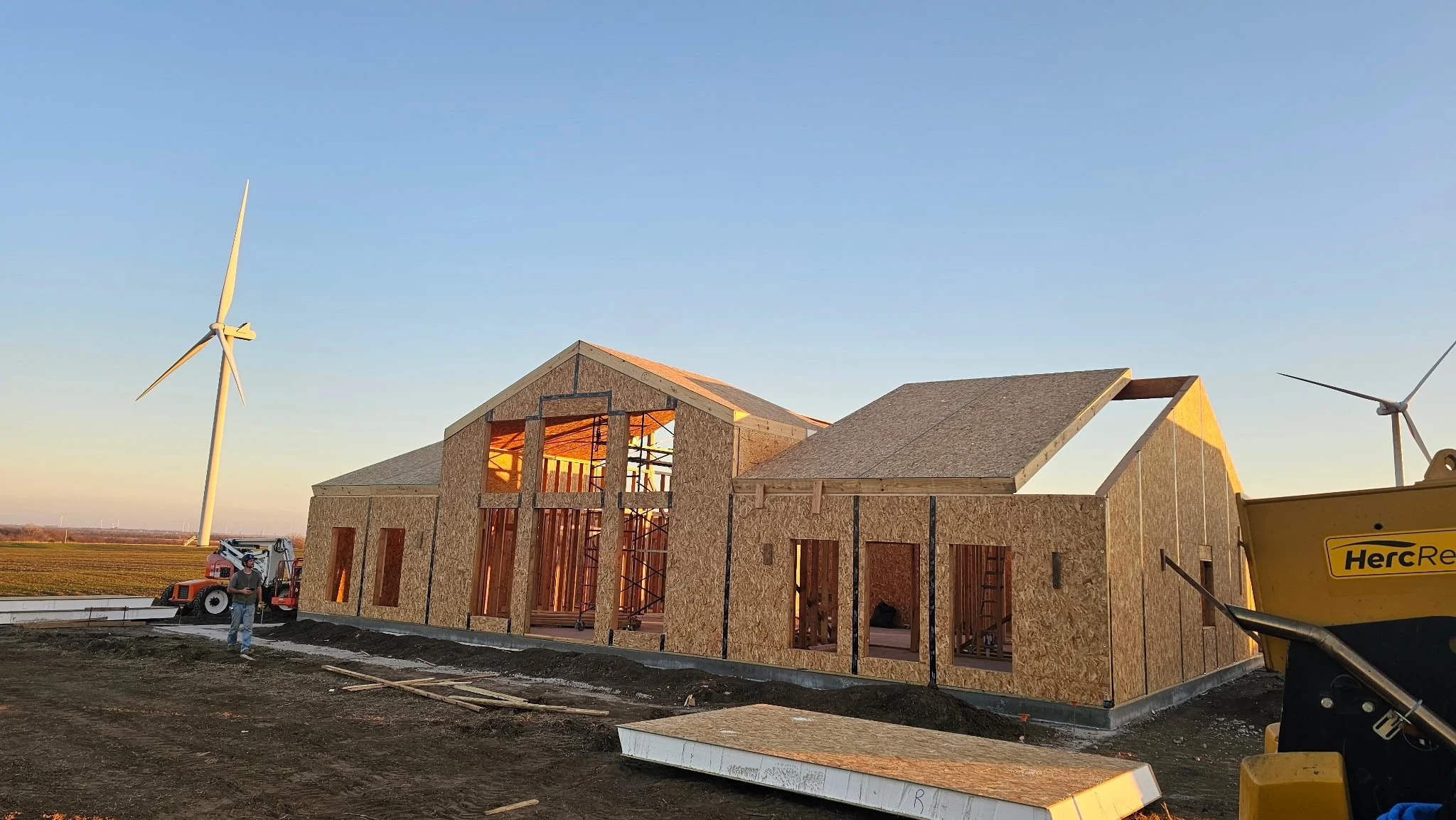 Construction site with a large wooden panel being lifted by a crane, adjacent to a partially completed building structure. A tractor is visible on the ground, surrounded by trees.