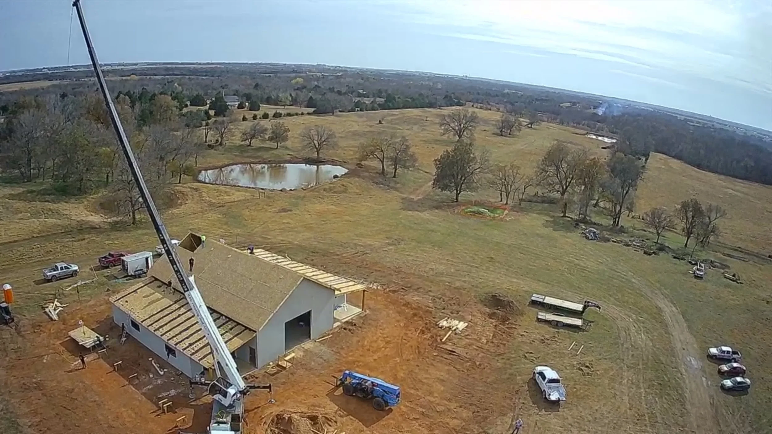 Construction site with a large wooden panel being lifted by a crane, adjacent to a partially completed building structure. A tractor is visible on the ground, surrounded by trees.