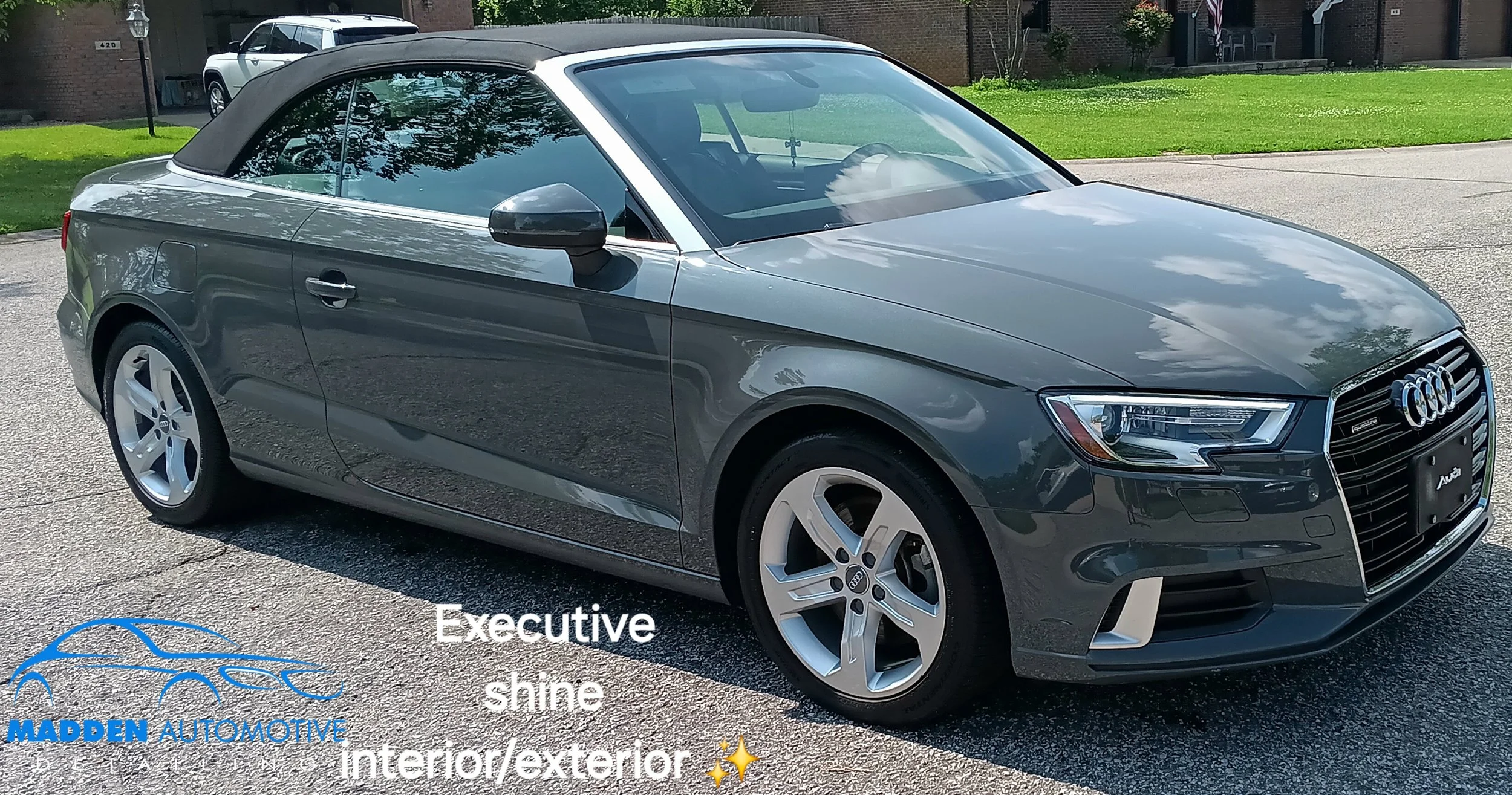 Gray Audi convertible with a black soft top parked outdoors on a driveway, reflecting clouds and trees on its surface.
