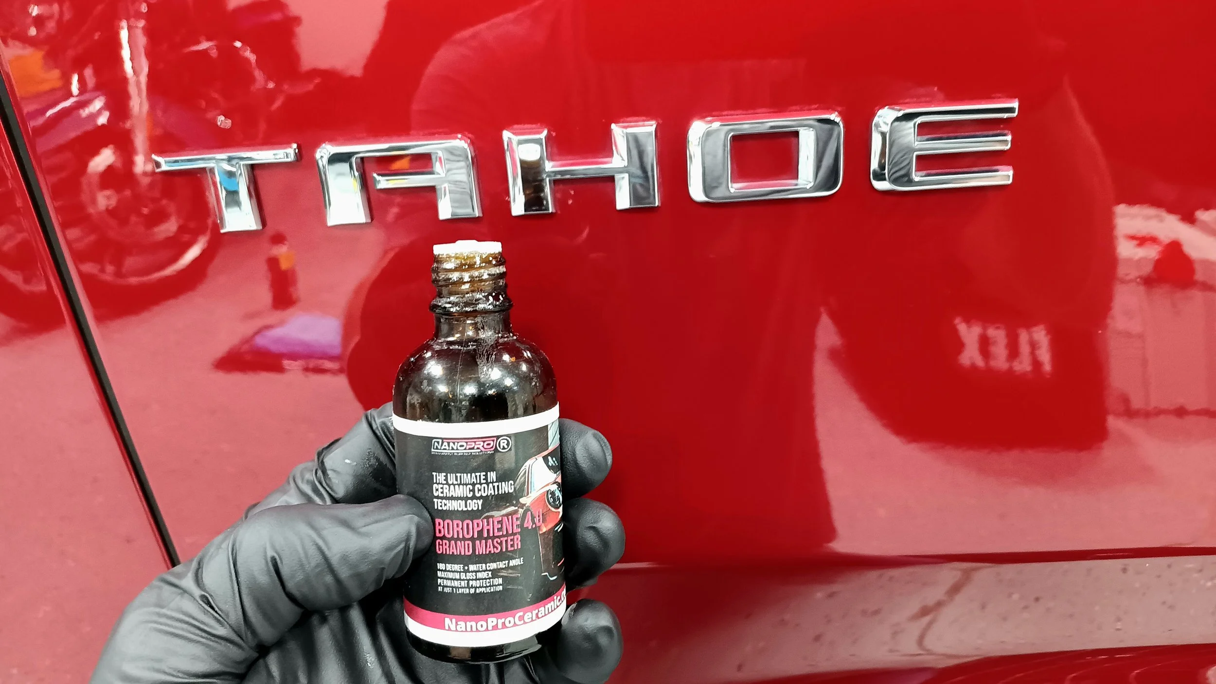 A person wearing black gloves holding a bottle of NanoPro Ceramic Borphen 4.0 Grand Master coating in front of a red car with a chrome 'THOR' badge