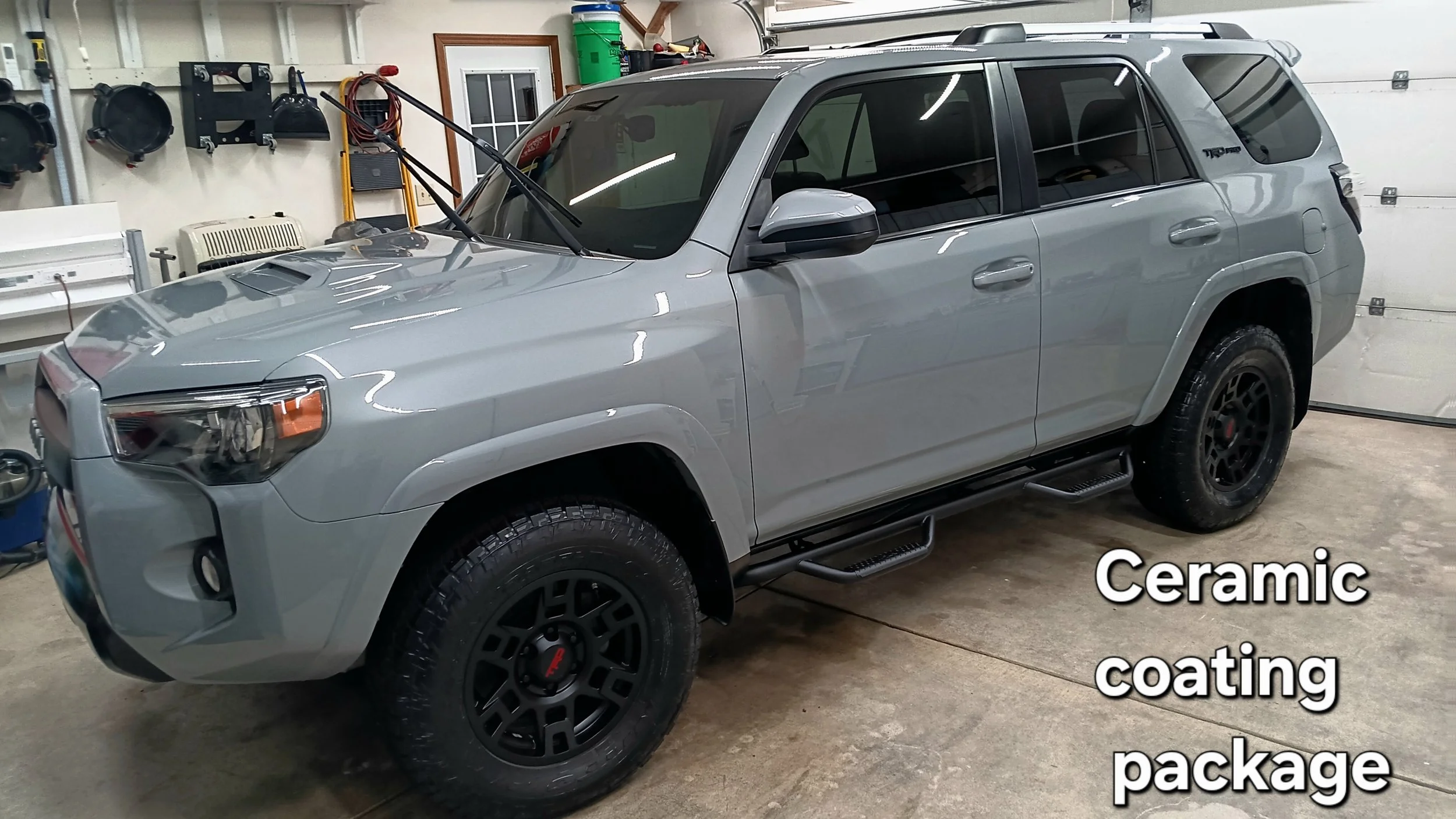 A gray SUV inside a garage with black wheels and side steps. The garage has tools and equipment on the wall, a green bucket, and a ladder. The text overlay highlights the vehicle's ceramic coating and packaging.