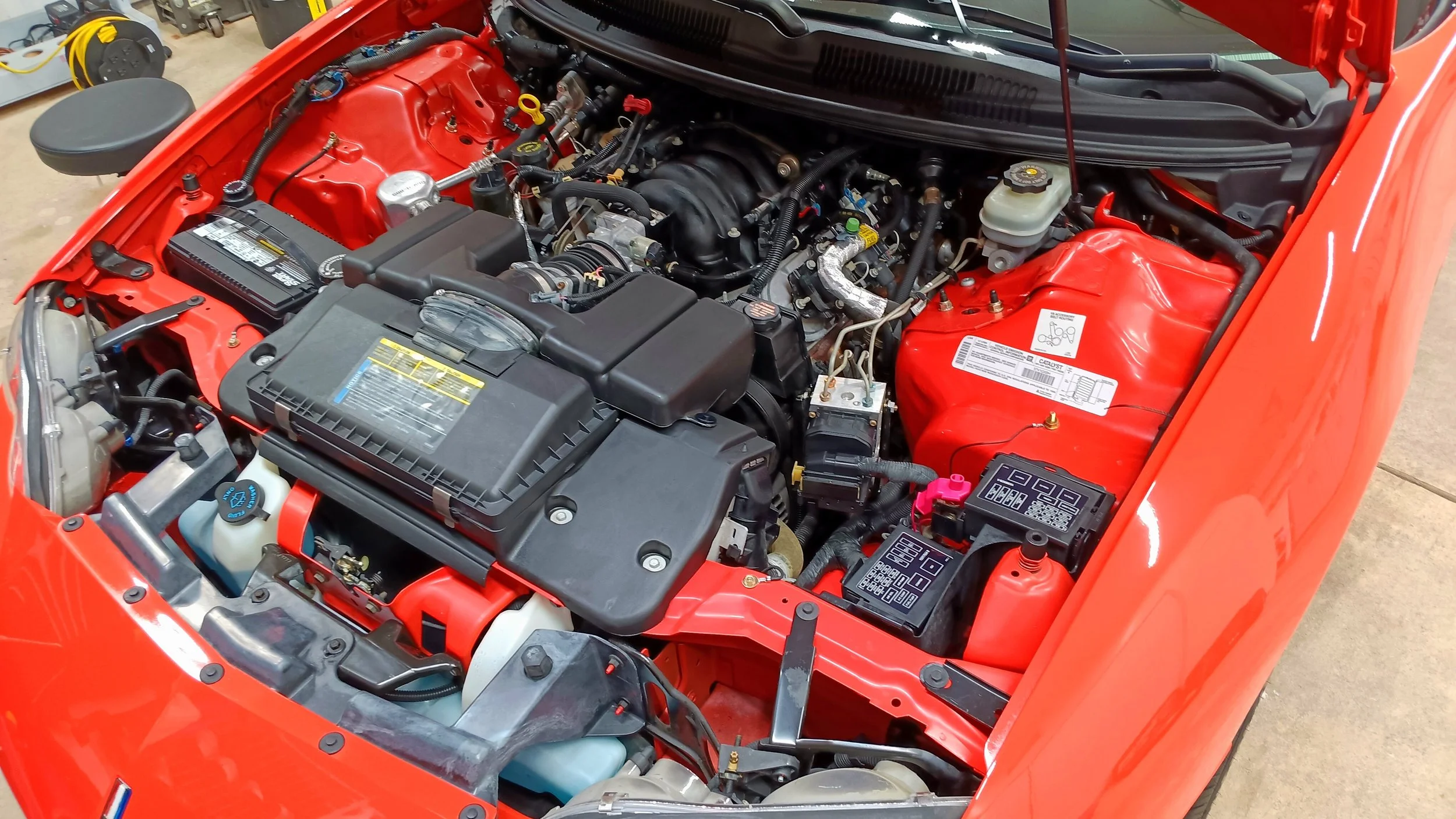 View of a car engine compartment with red painted body, black engine components, and various wires, hoses, and containers, indicating a clean and organized engine bay.