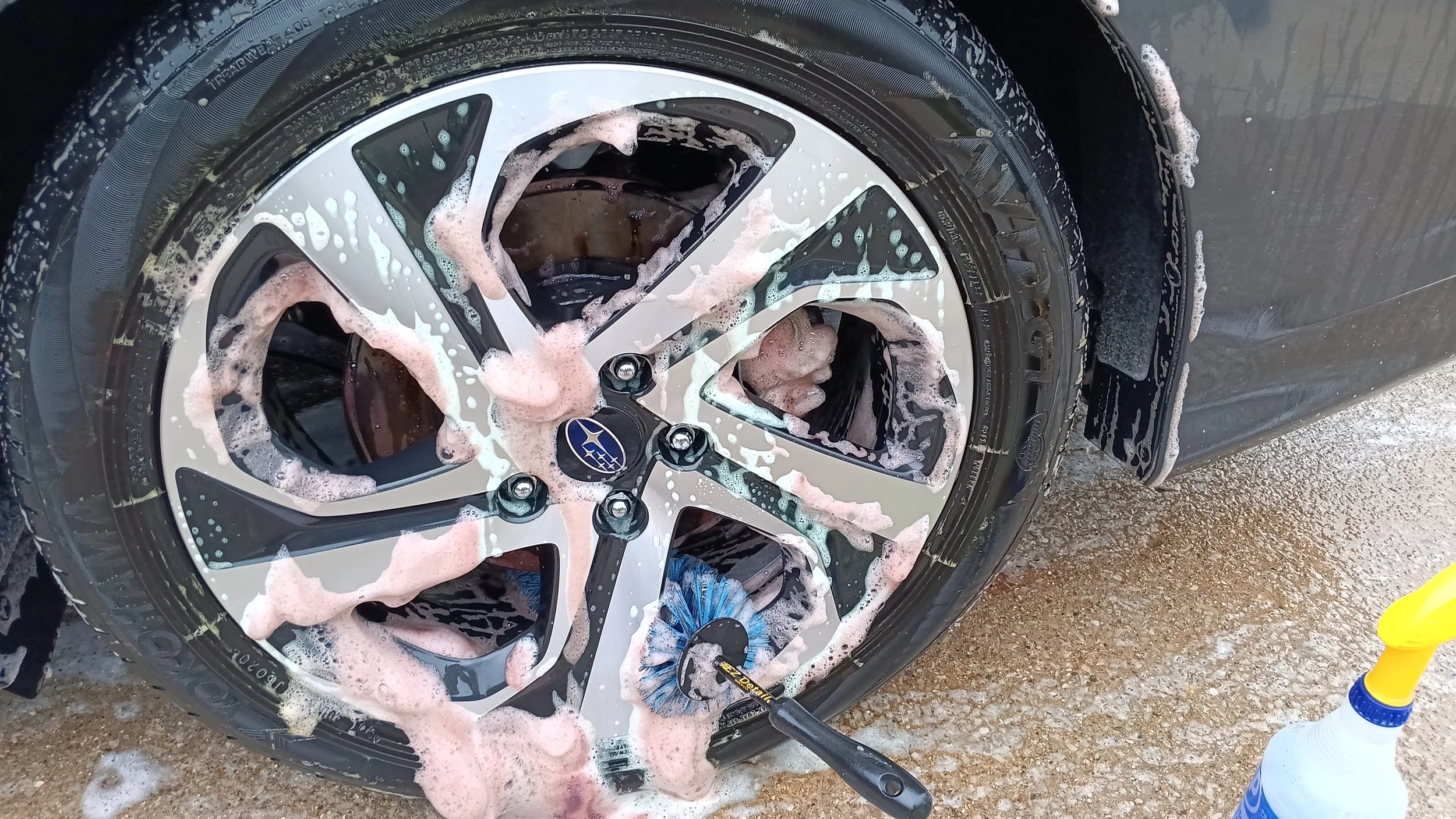 A car tire with a Subaru logo being cleaned with soap and a scrub brush, with soap suds and water on the tire and the ground.