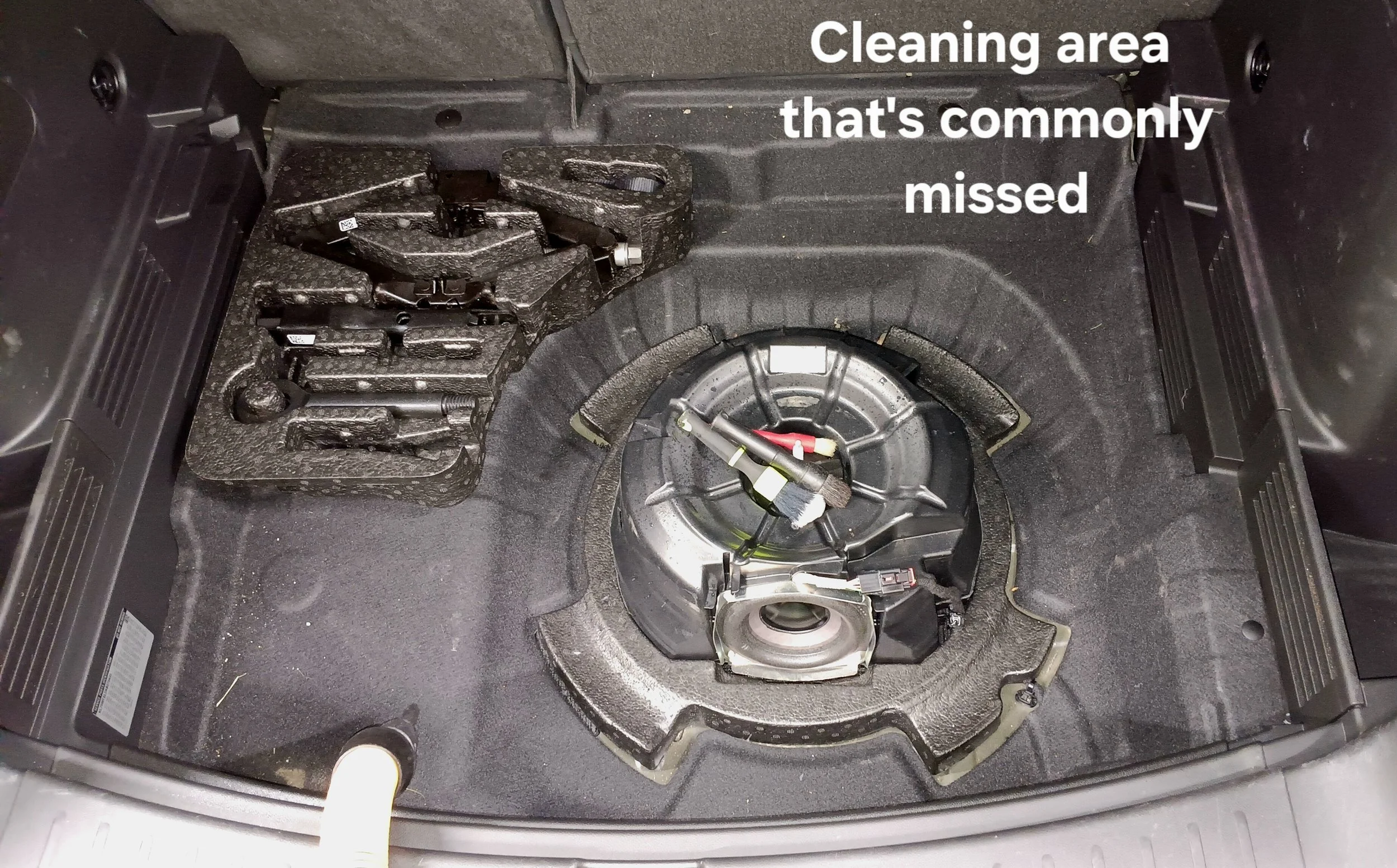 The trunk of a car with a removed spare tire, exposing the empty space with foam tools to the left and a circular metal mounting area in the center, with a caption reading 'Cleaning area that's commonly missed'.
