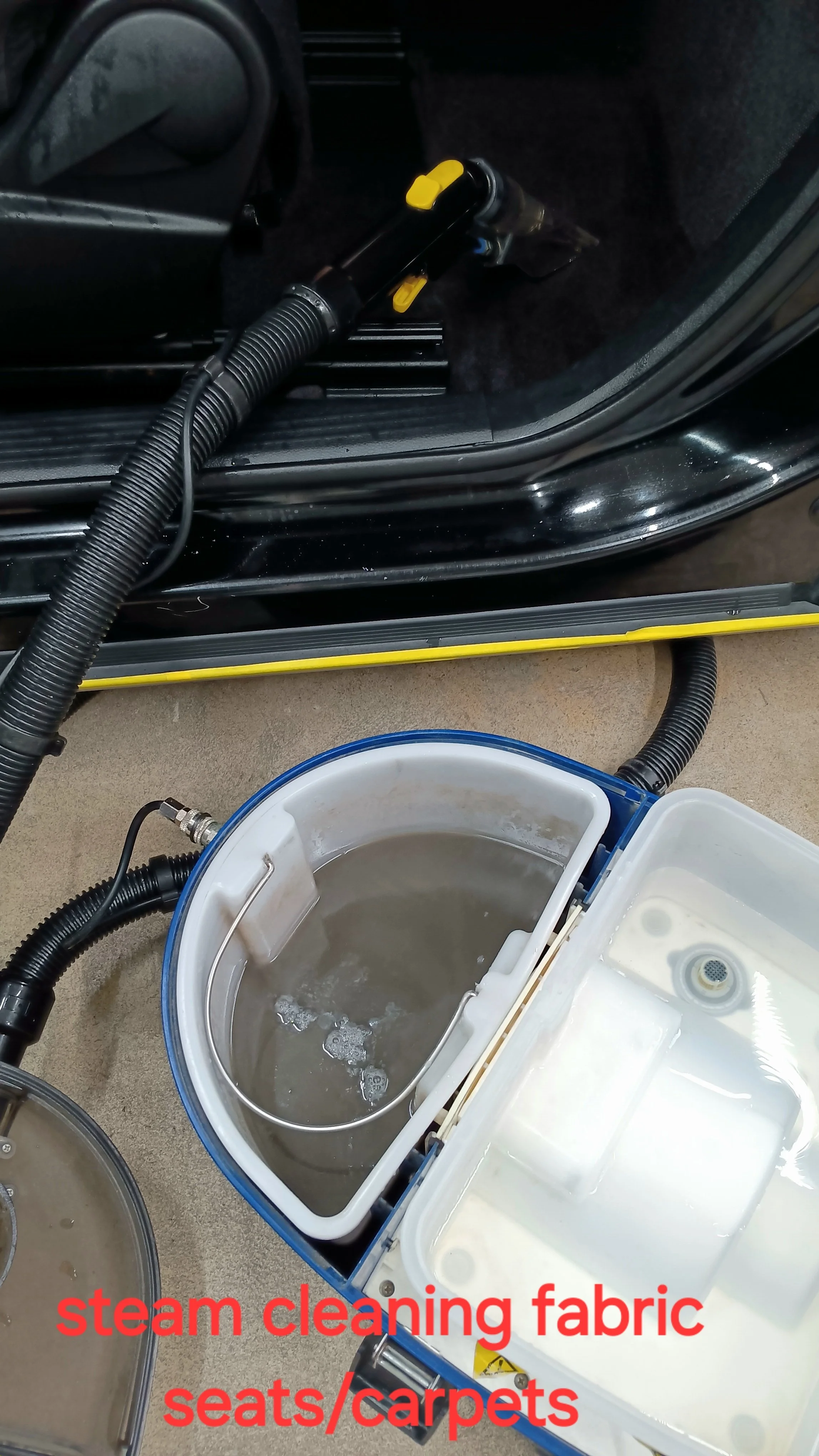 A steam cleaning machine with water inside, connected to a vehicle interior with vacuum hoses, used for cleaning fabric seats and carpets.