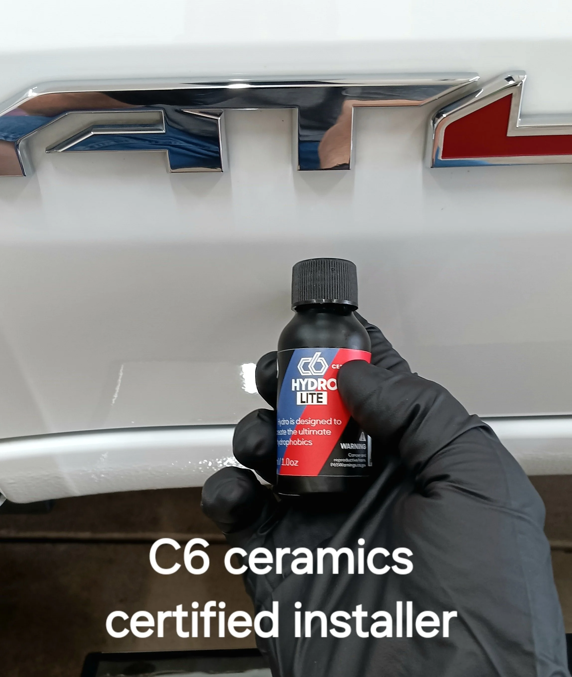 A person wearing a black glove holding a bottle of HydroSilex HydroLITE ceramic coating in front of a white car with a C6 Corvette emblem. The text overlaid on the image says, 'C6 ceramics certified installer'.