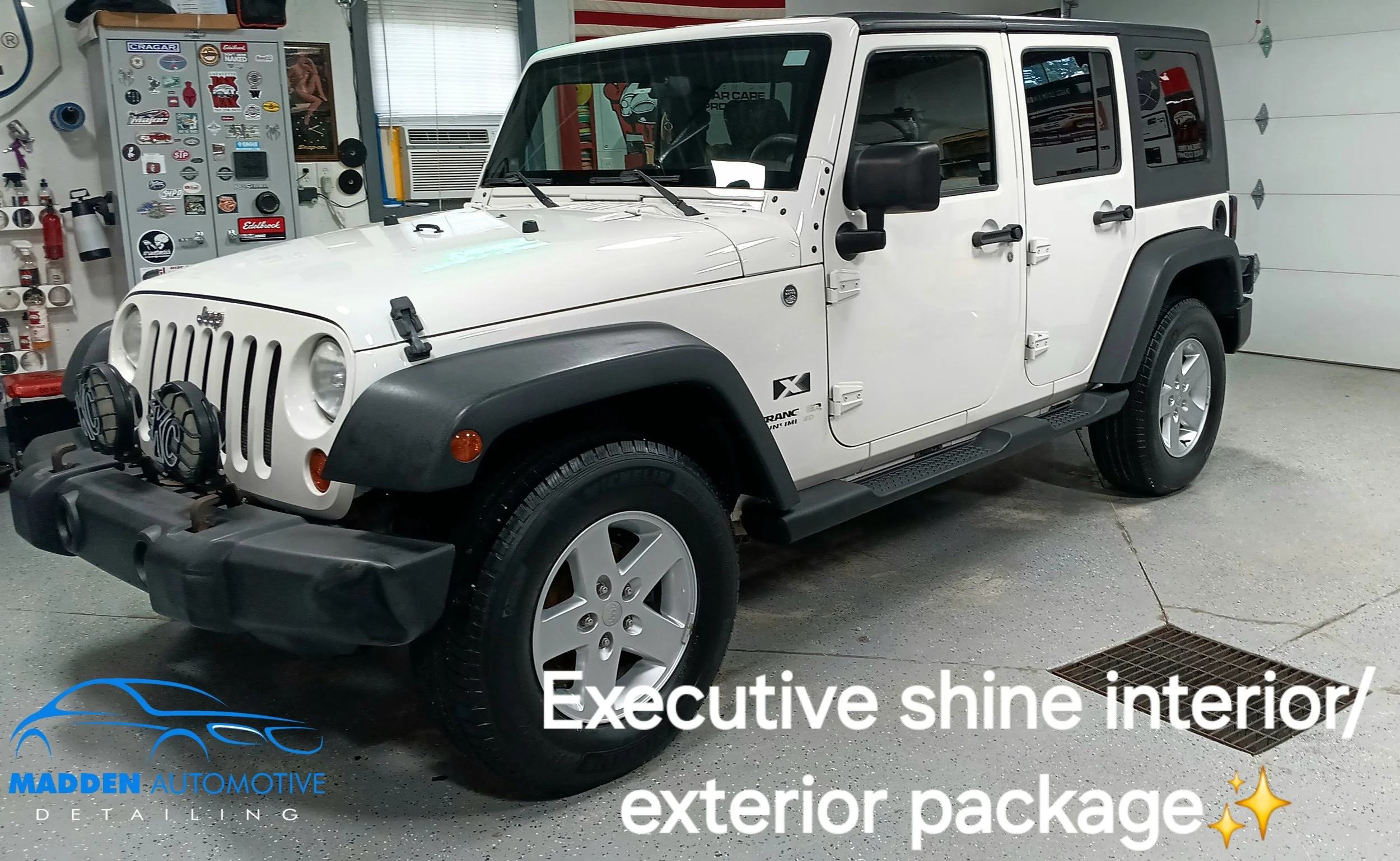 White Jeep Wrangler parked indoors on a gray concrete floor, with black fender flares and two front black grille-mounted auxiliary lights. The background includes garage shelves with car accessories and a closed garage door.