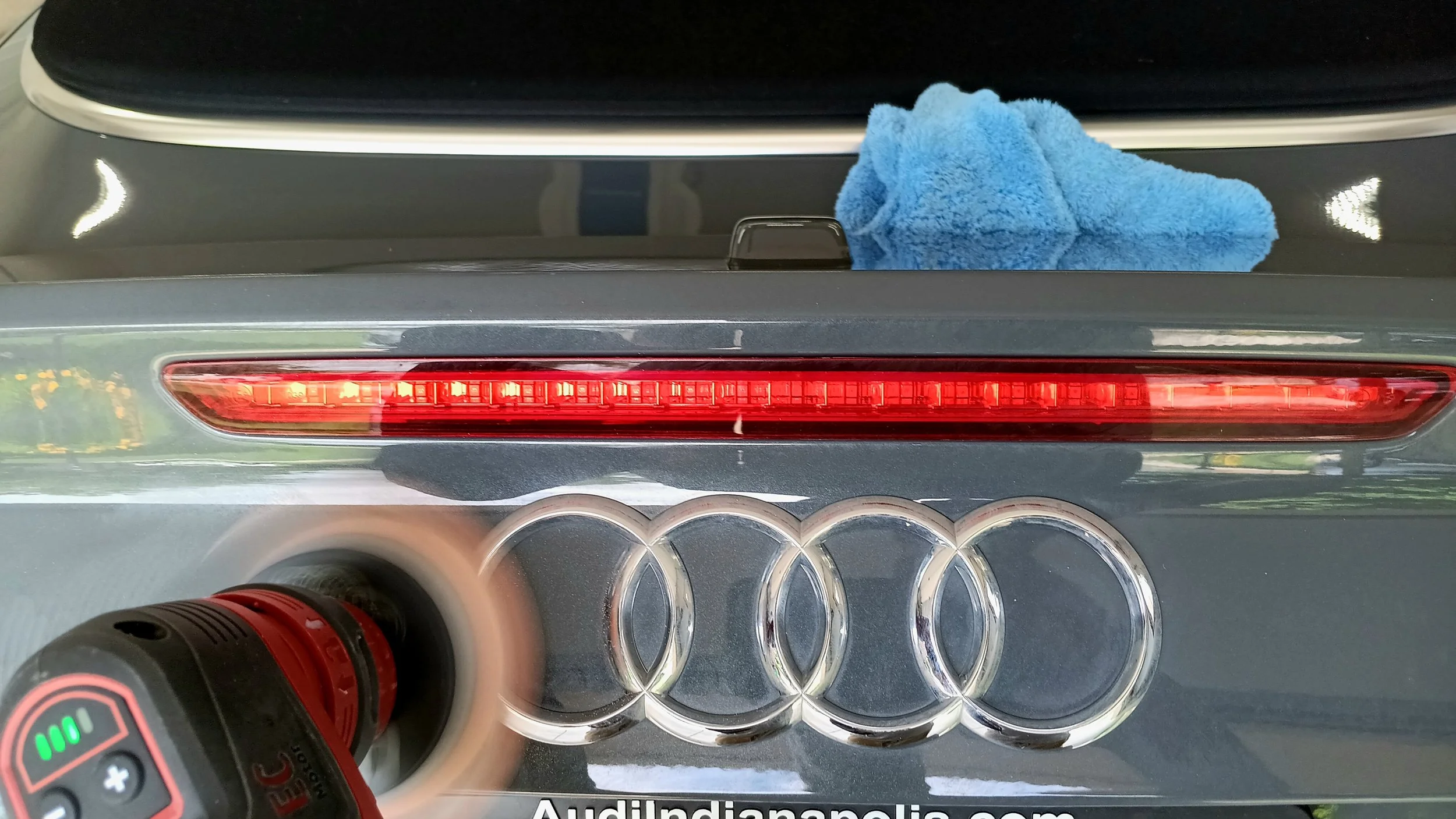 Car trunk with four Audi rings emblem, a red LED tail light, a blue microfiber cloth on the black interior, and a polishing machine in motion.