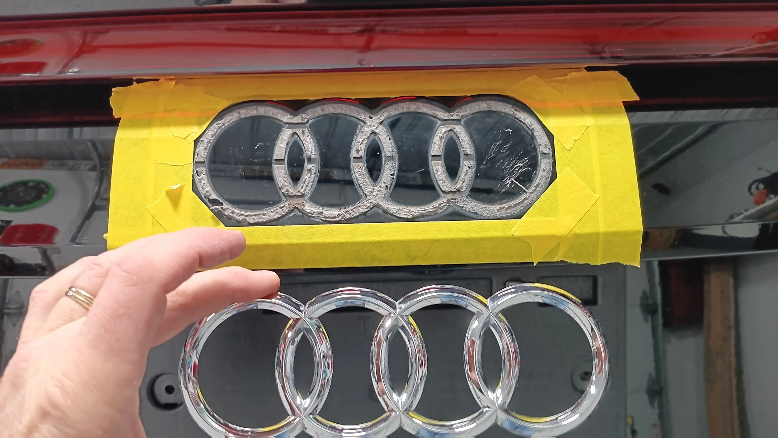 Someone holding a clear Audi logo made of glass or acrylic, with a set of five smaller clear logo rings below, in front of a black surface with a taped rectangular cutout holding a metallic emblem of five interlinked rings.