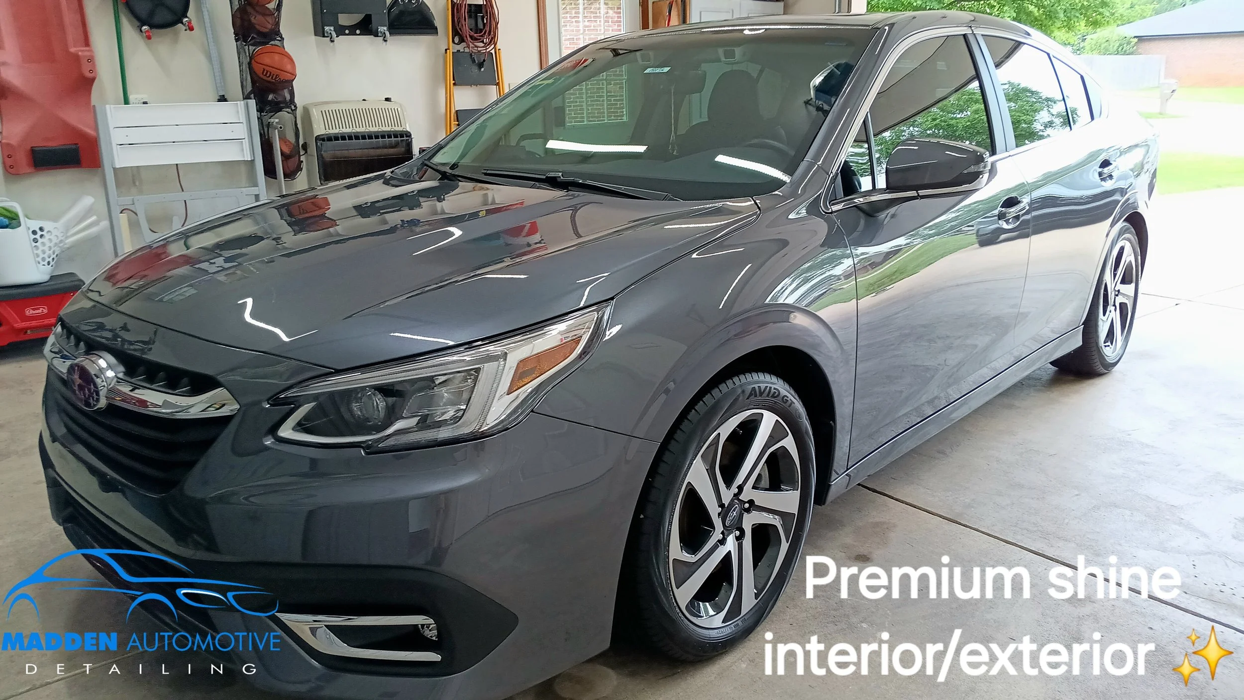 A grey sedan parked inside a garage with a shiny exterior. The garage has shelves and various tools in the background. The car's surface reflects light, indicating a premium shine, with the text "Premium shine interior/exterior" and a logo for Madden
