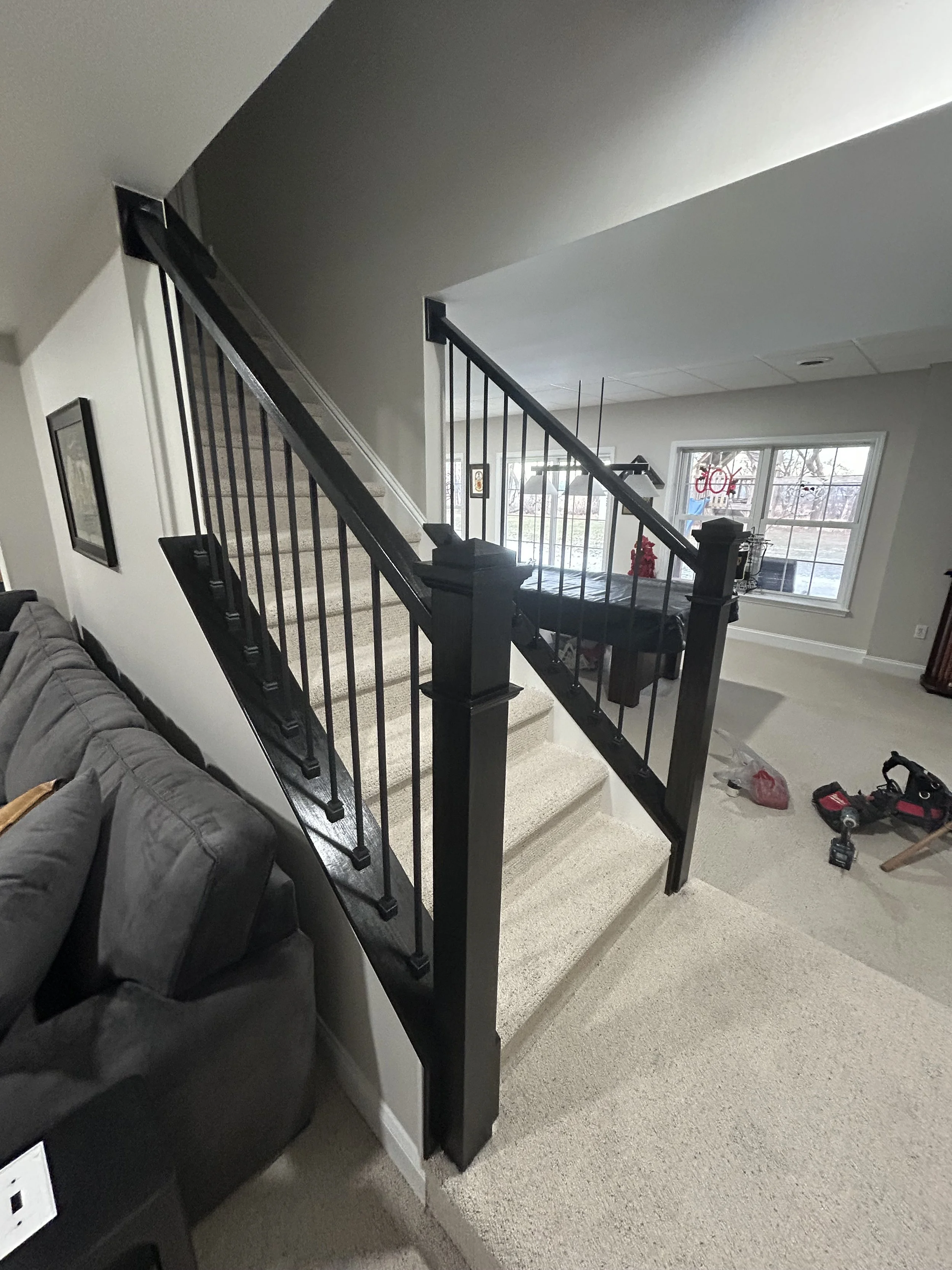 Interior view of a house showing stairs with black railing, carpeted steps, and a living room area with furniture and tools, large window with a view of the outdoors.