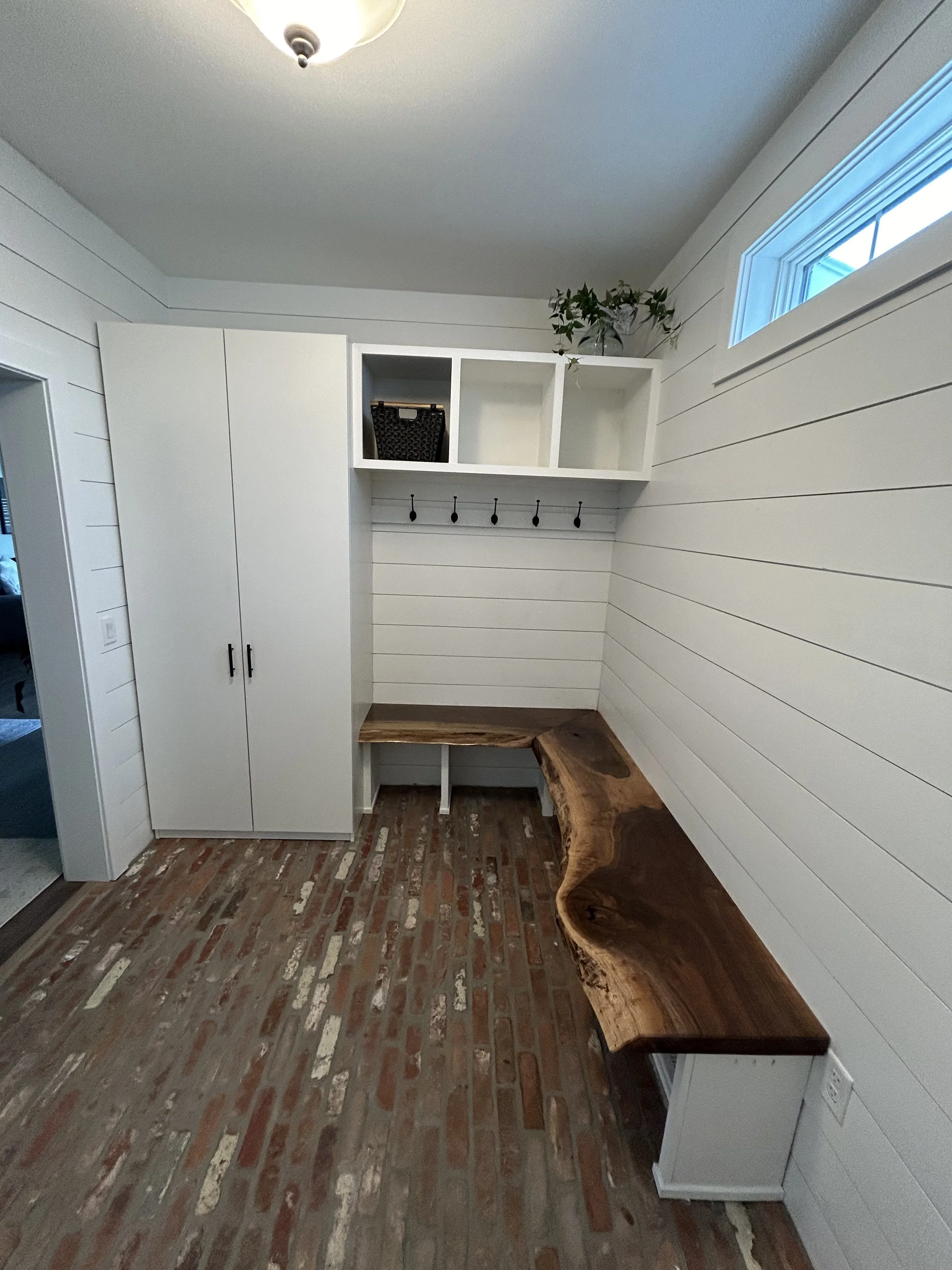 A mudroom with white shiplap walls, a wooden bench with a natural edge, white cabinetry, and open cubbies. There are black hooks on the wall below the cubbies, a small window, and a light fixture on the ceiling.