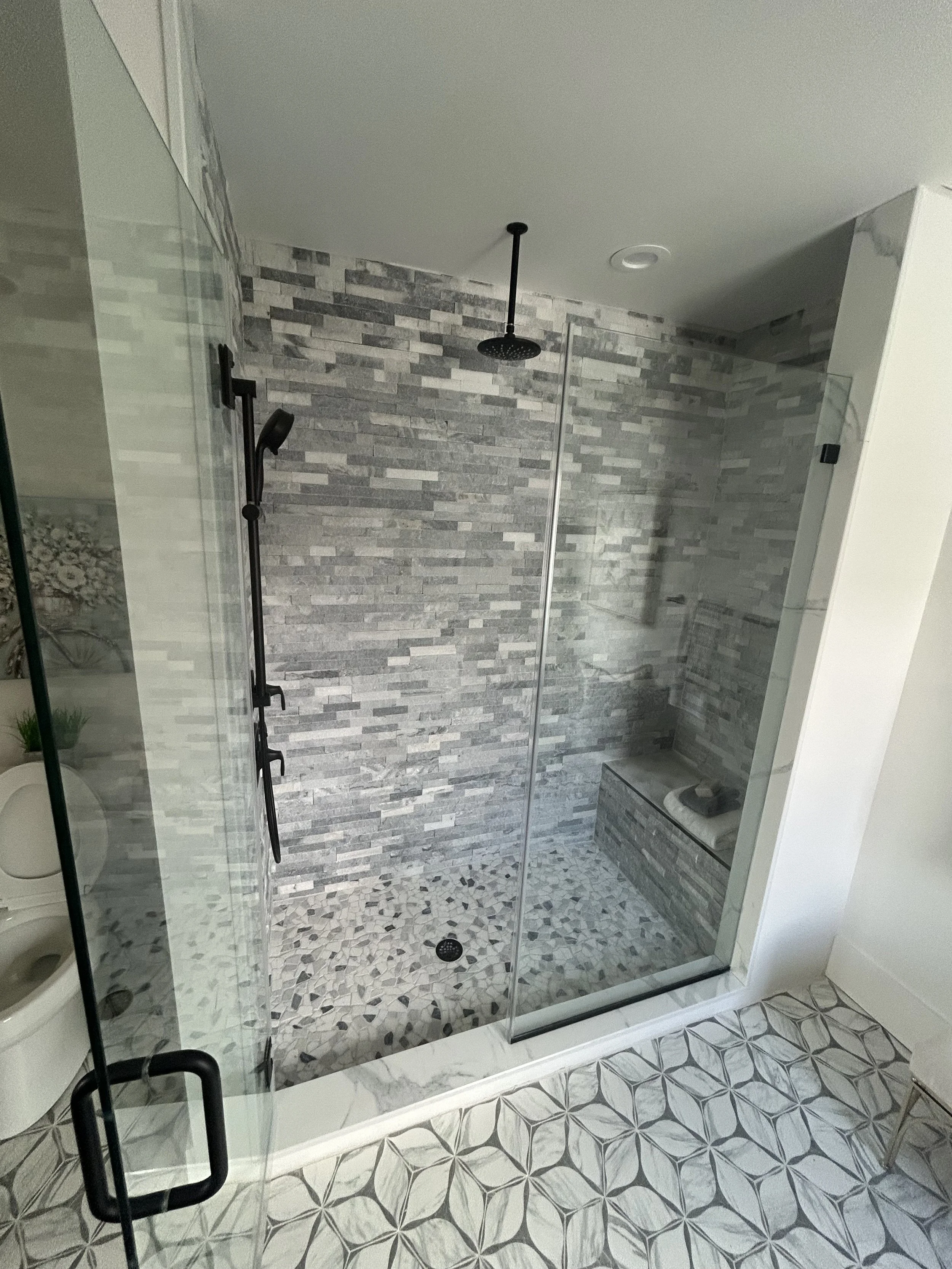 Modern walk-in shower with gray stone wall tiles, pebble floor, a ceiling-mounted rainfall showerhead, a black handheld shower, and a built-in bench in a bathroom with patterned tile flooring.