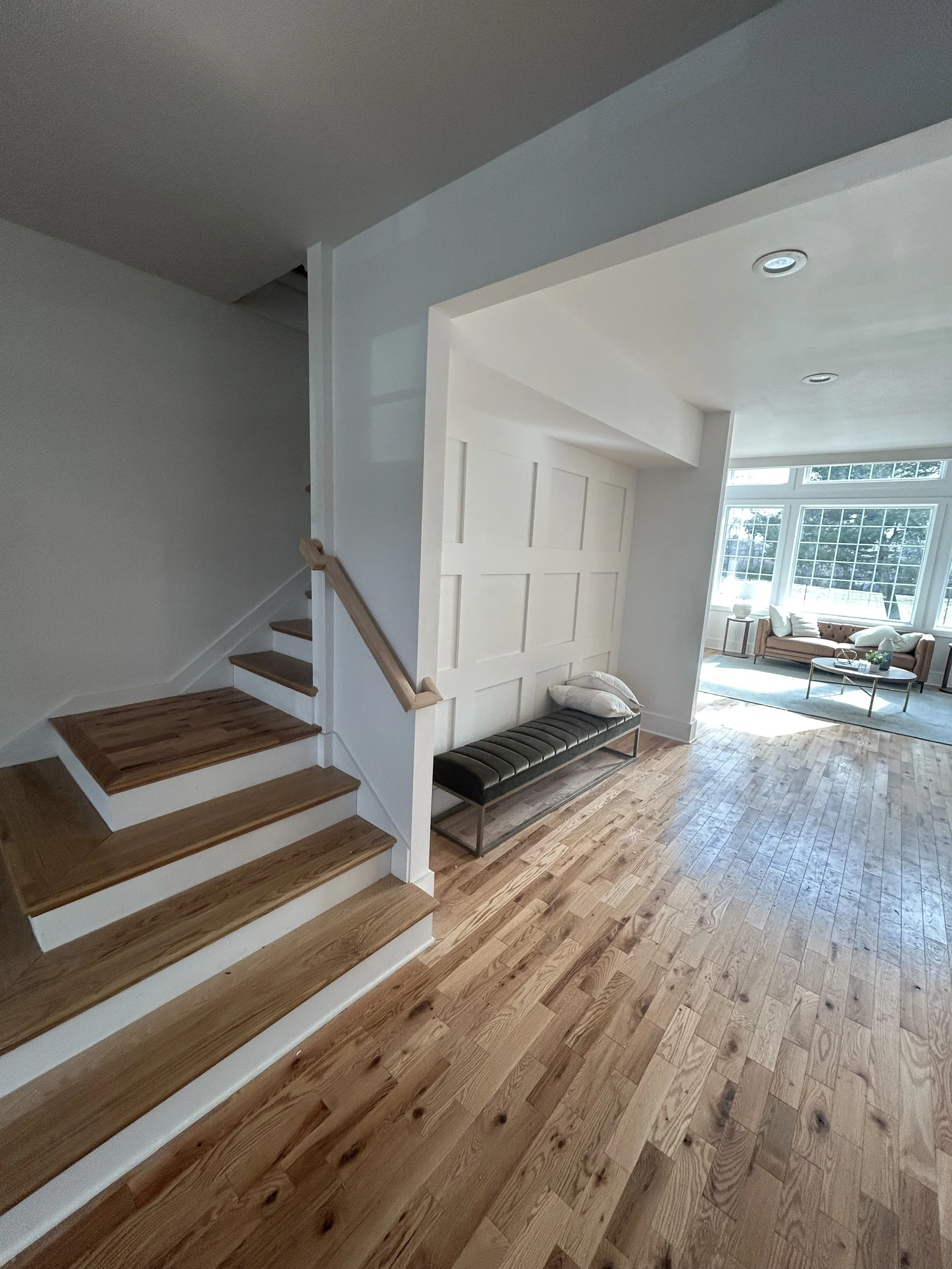 Living room with hardwood floors, a staircase with wooden steps, white walls, large windows, and a couch with pillows.