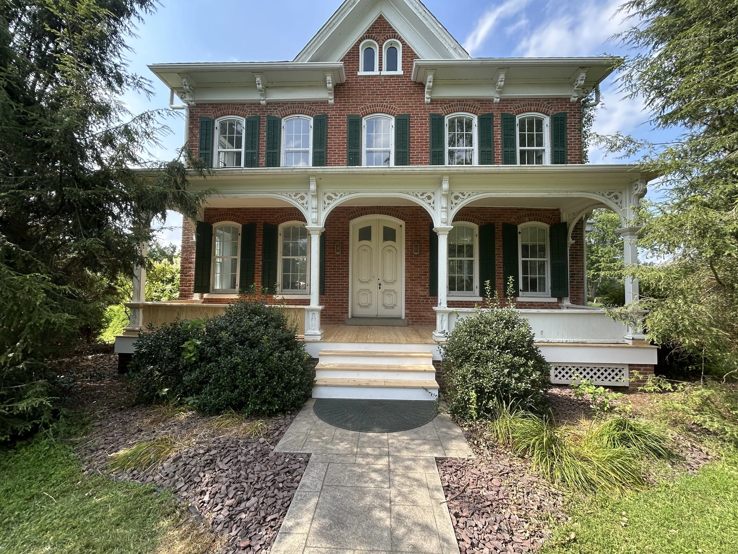 A large, historic brick house with a front porch, white decorative trim, dark green shutters, and multiple windows, surrounded by greenery and trees.