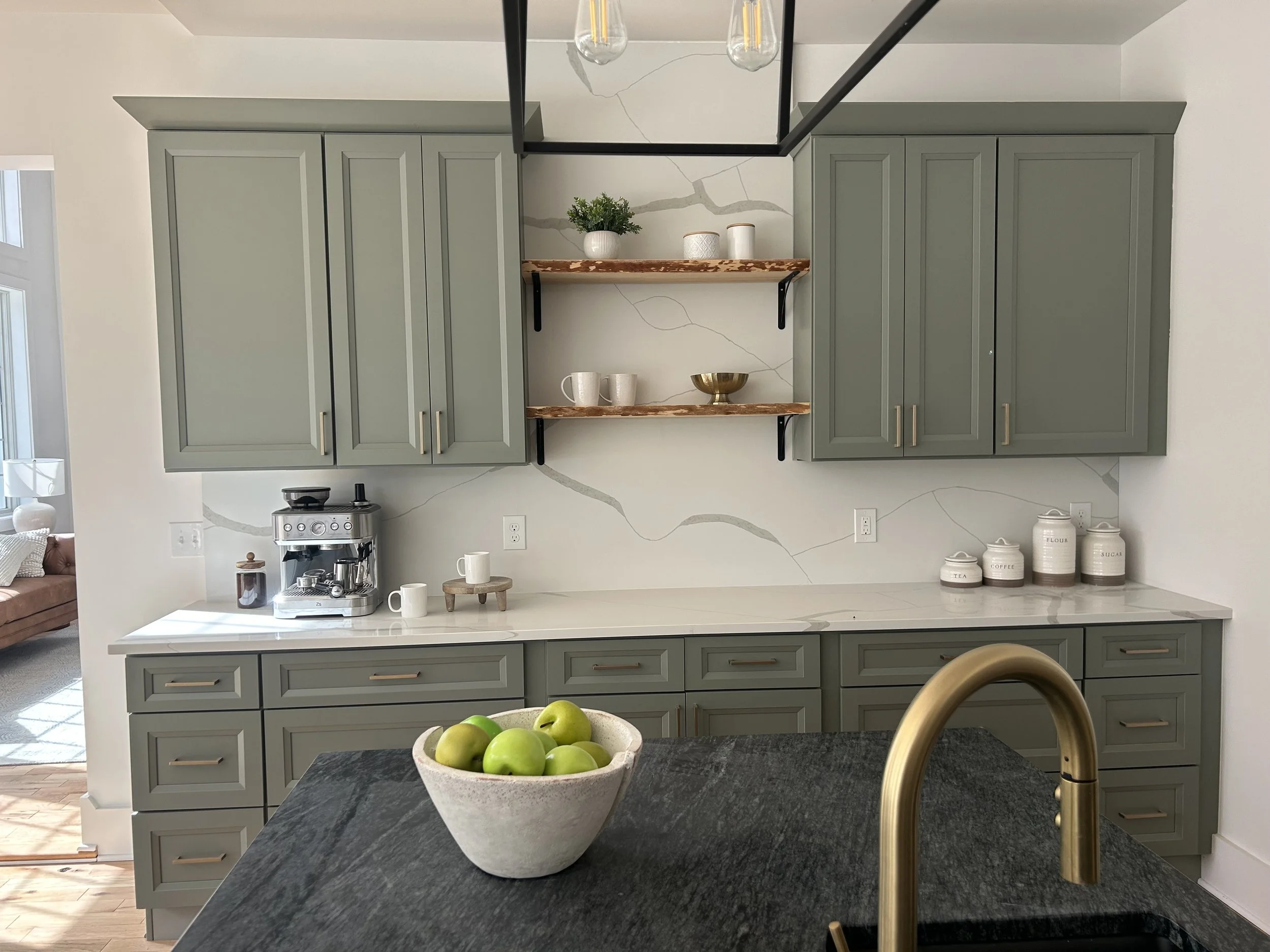 Kitchen with green cabinets, white marble backsplash, open wooden shelves, a countertop with a coffee machine, cups, and jars, and a black countertop island with a bowl of green apples and a gold faucet.