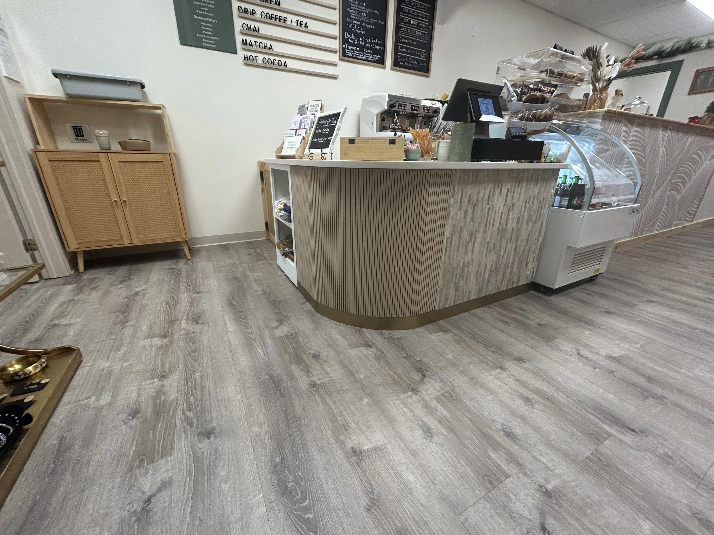 Coffee shop counter with a cash register, display case with baked goods, and a menu on the wall listing drinks like drip coffee, tea, chai, matcha, and hot cocoa.