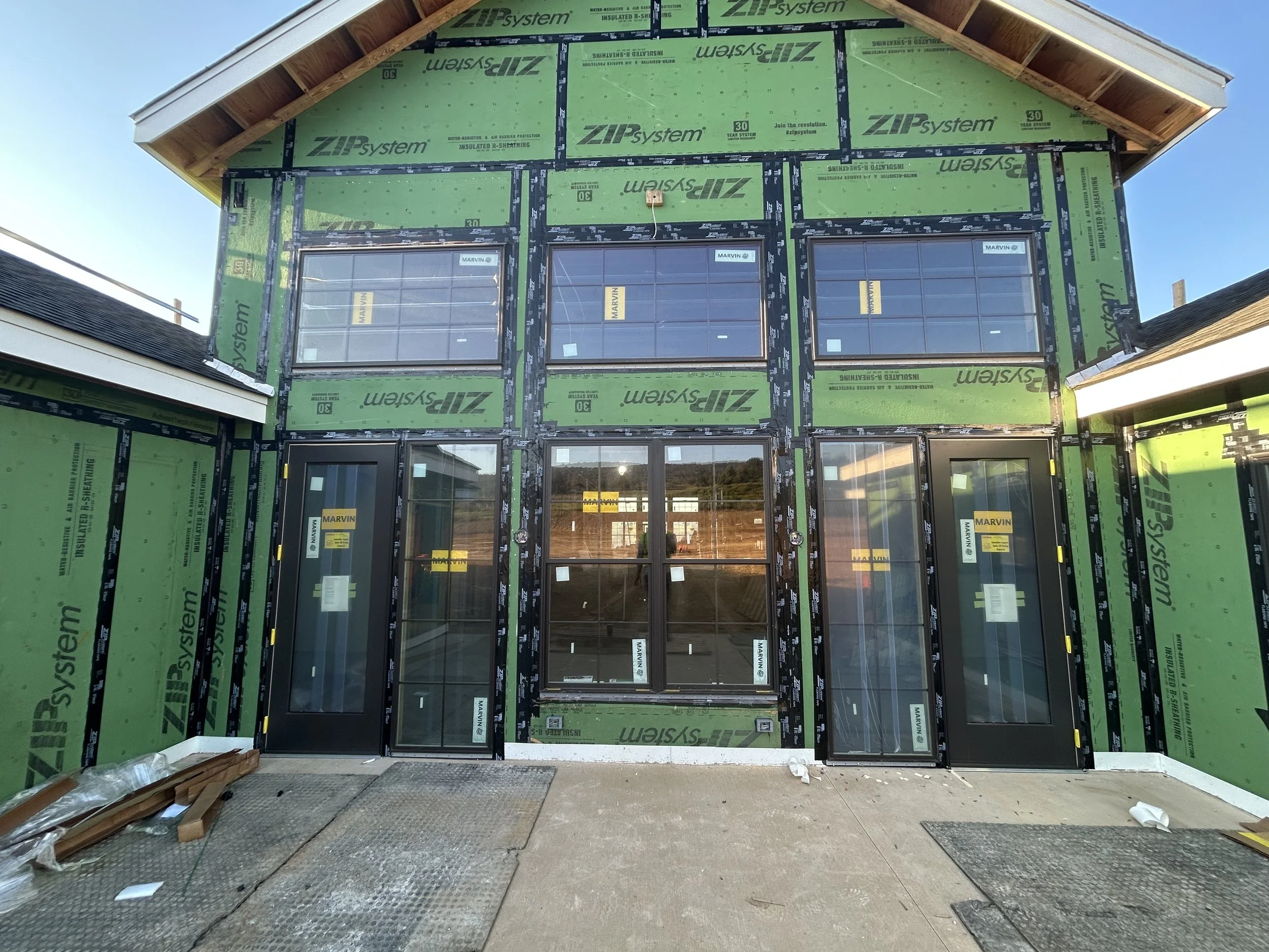 Facade of a house under construction with green insulation and black-framed sliding glass doors and windows.