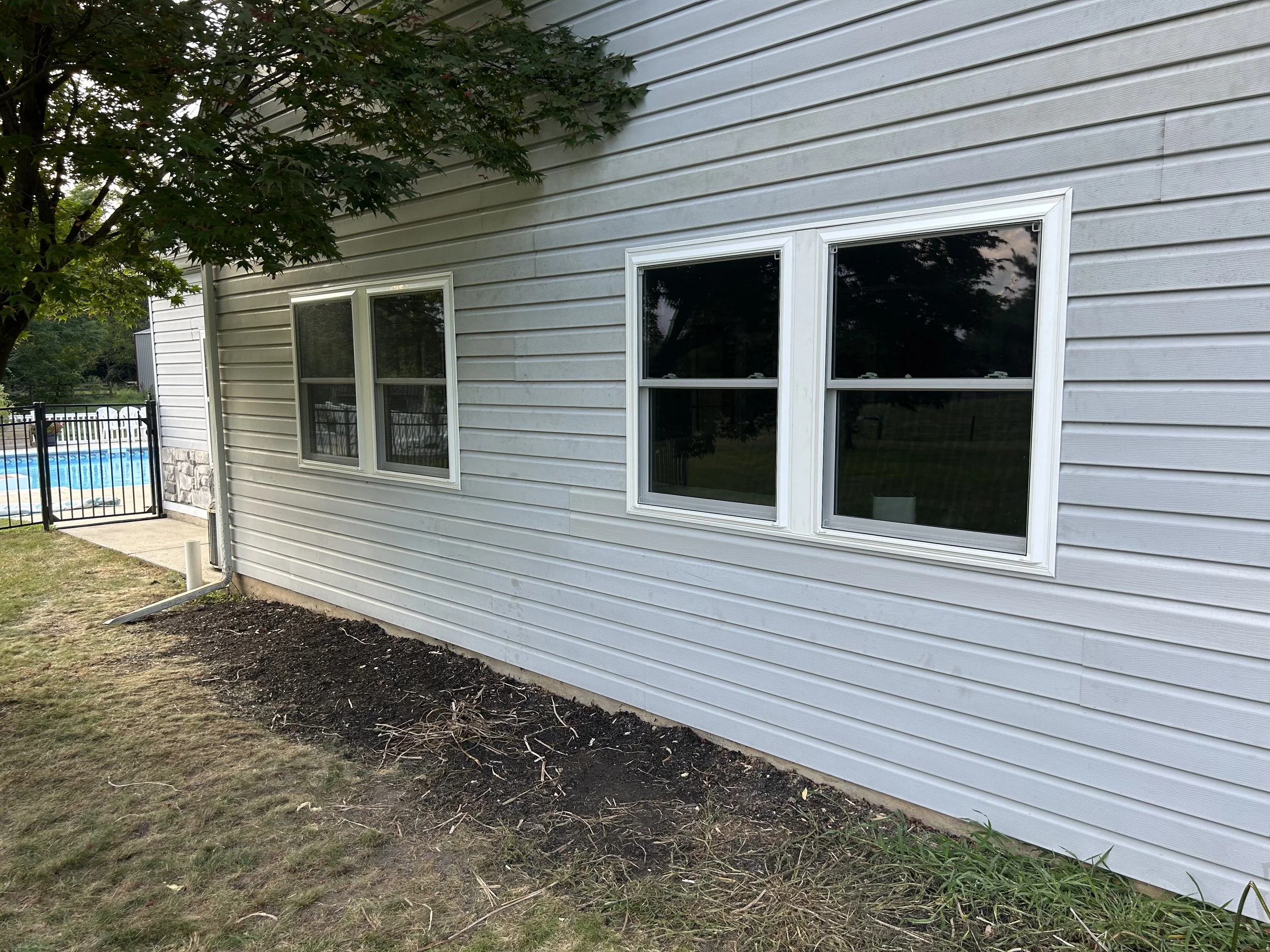 Back of a house with white siding and two white-framed windows, a tree with green leaves, and a garden bed with soil in front of the house. A swimming pool with a black fence is visible in the background.