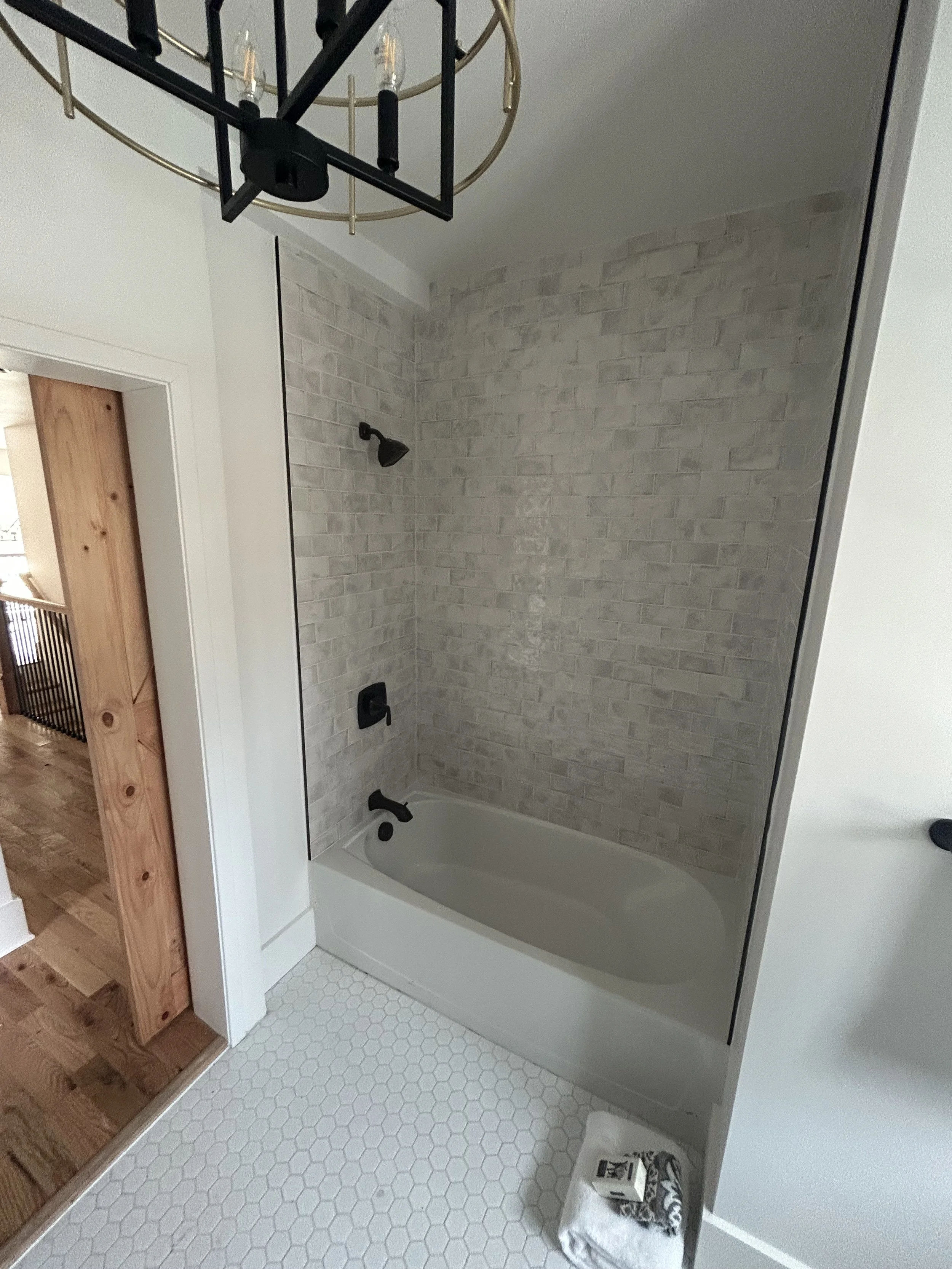 Bathroom with a bathtub, gray brick tile wall, black fixtures, white hexagon floor tiles, and a chandelier overhead