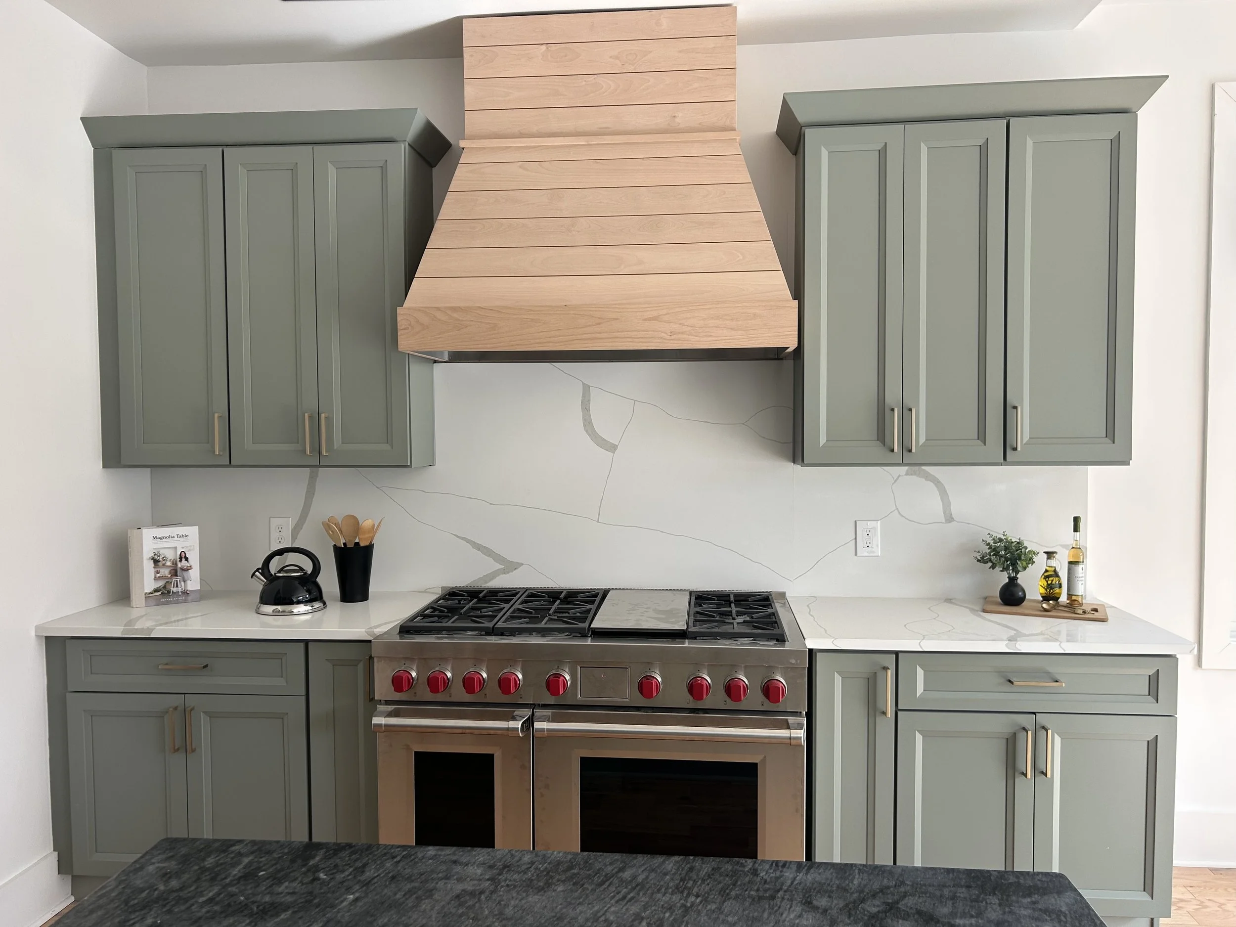 Kitchen with light green cabinets, a stainless steel stove with red knobs, a wooden range hood, marble backsplash, and a white marble countertop with a small potted plant and bottles on the right side.