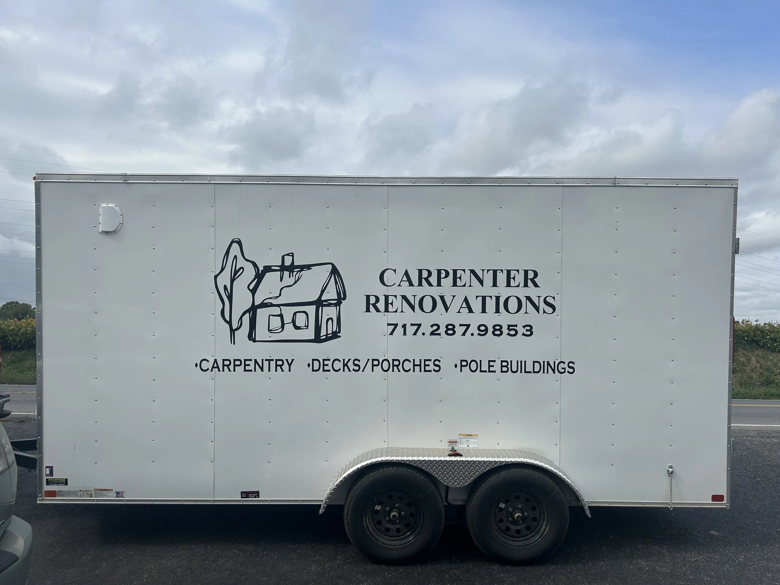 White trailer with logo and text for Carpenter Renovations, featuring a line drawing of a house and tree, and the company's phone number and services such as carpentry, decks/porches, and pole buildings