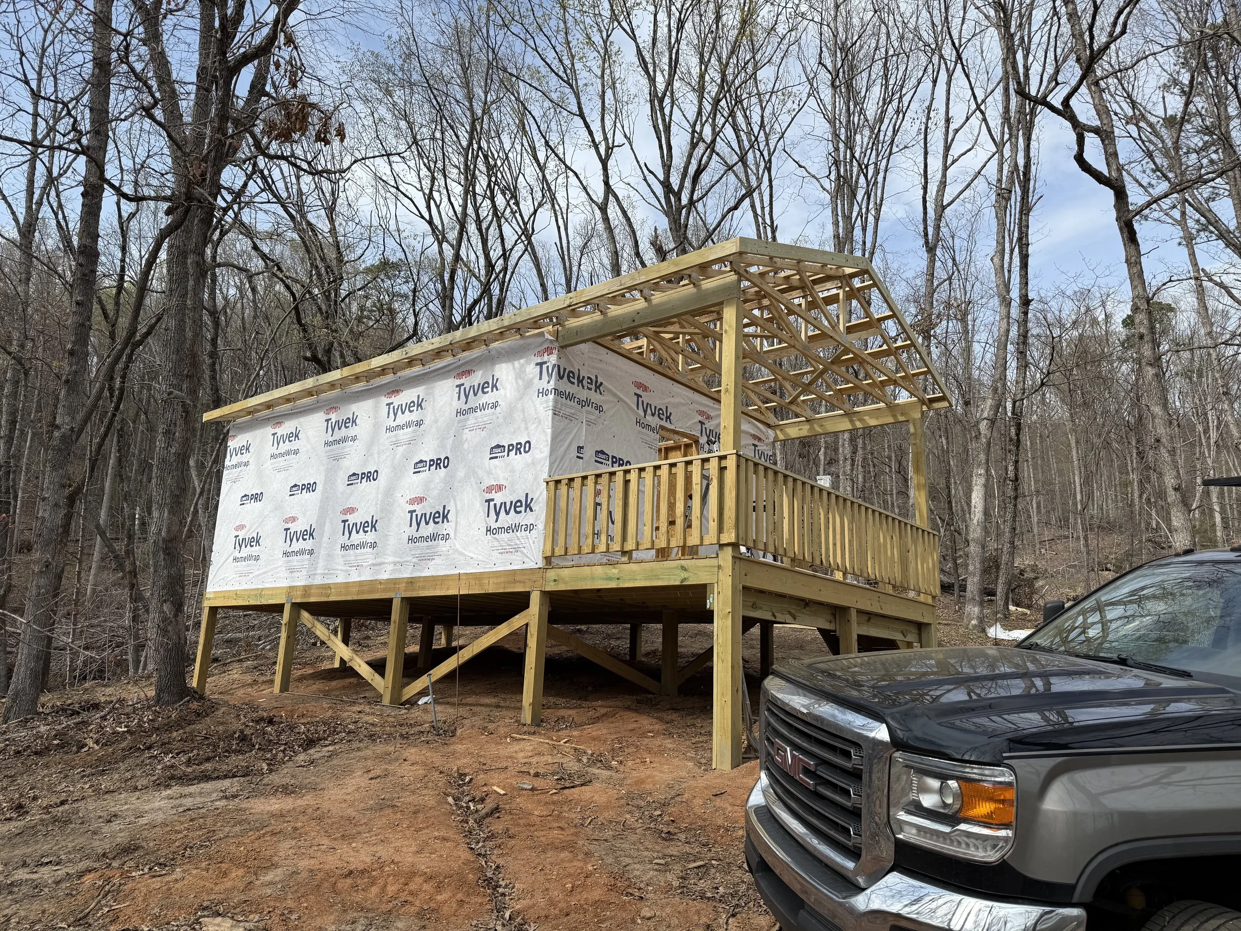 Under construction house on a wooded lot with a front porch, partially covered roof, wrapped in Tyvek weather barrier, and a black GMC truck parked nearby.