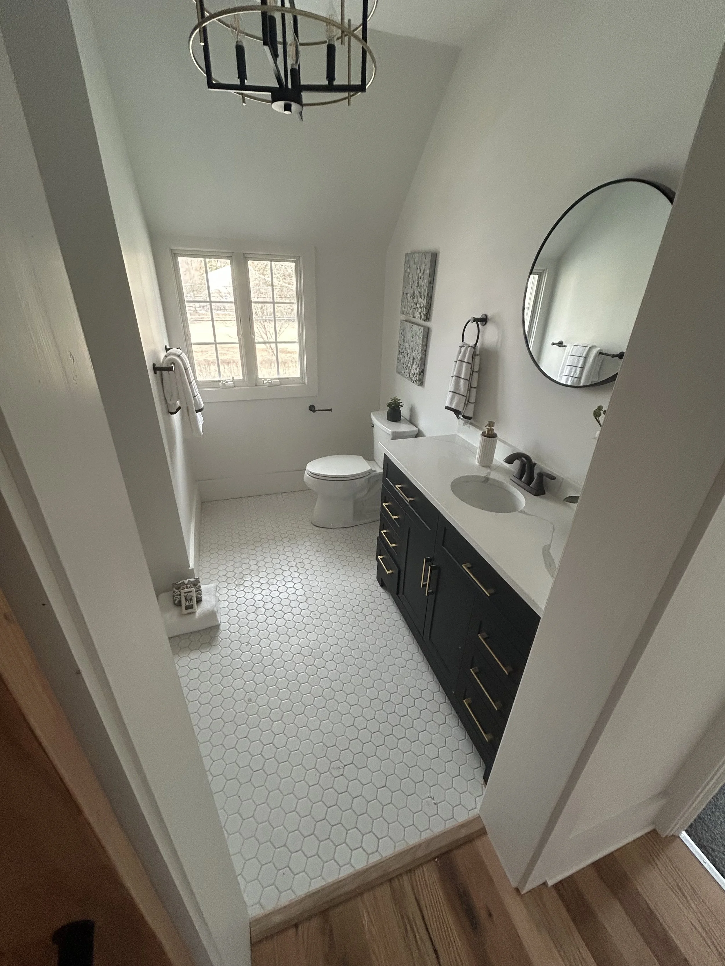 A modern bathroom with a white hexagon tile floor, black vanity with gold handles, oval mirror, white countertop, and a white toilet near a window with four panes, decorated with wall art and small plants.