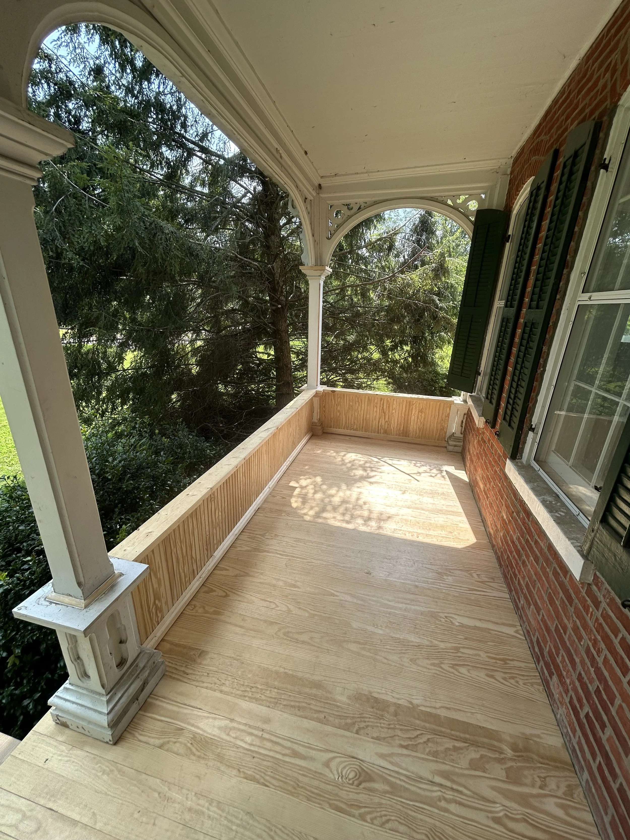 A front porch with new wooden flooring and green shutters on the brick wall, overlooking a tree-filled yard.