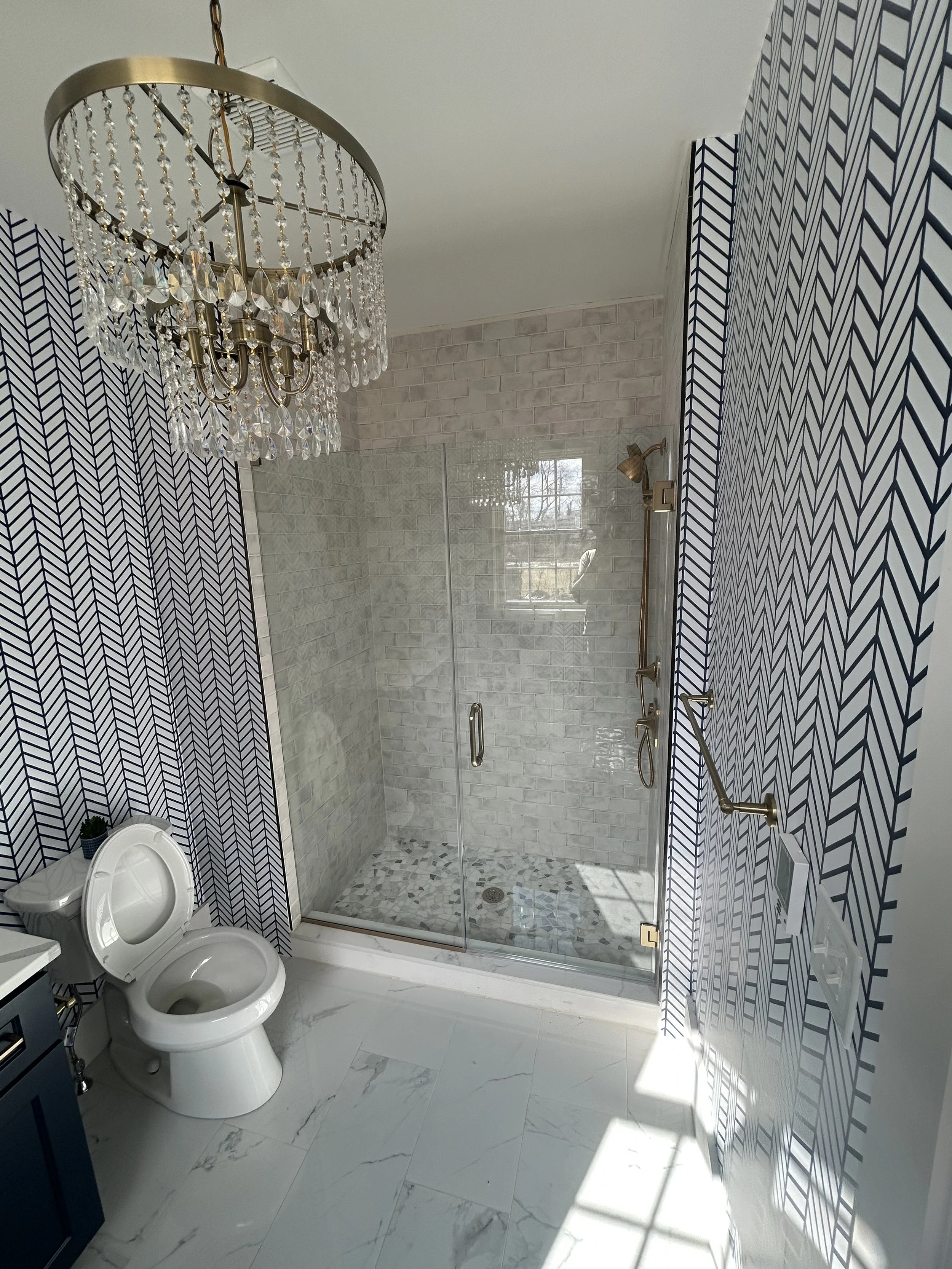 Bathroom with patterned black and white wallpaper, a crystal chandelier, a glass shower with white brick tiles, a white toilet, and marble-like flooring.
