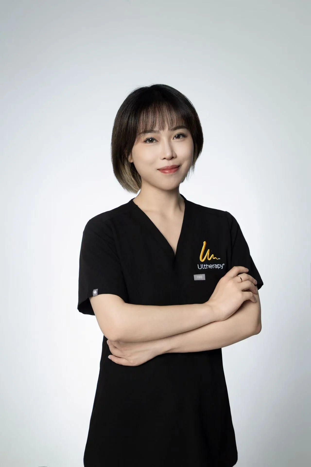 A woman in a black shirt with Ultherapy logo and yellow writing, crossing her arms and smiling against a plain gray background.