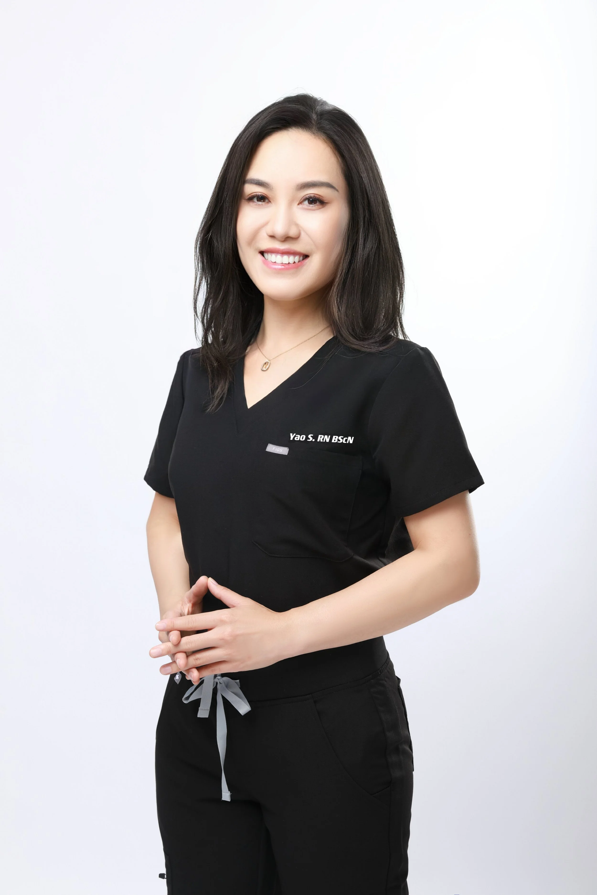 A woman with long black hair, wearing a black medical scrub top with her name badge, smiling and standing against a plain white background.