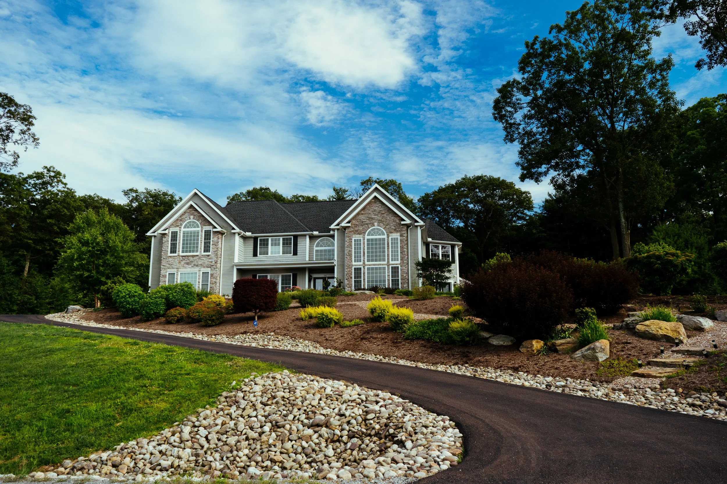 A large, two-story house with a manicured front yard and a curved driveway, set against a background of green trees and a partly cloudy blue sky.
