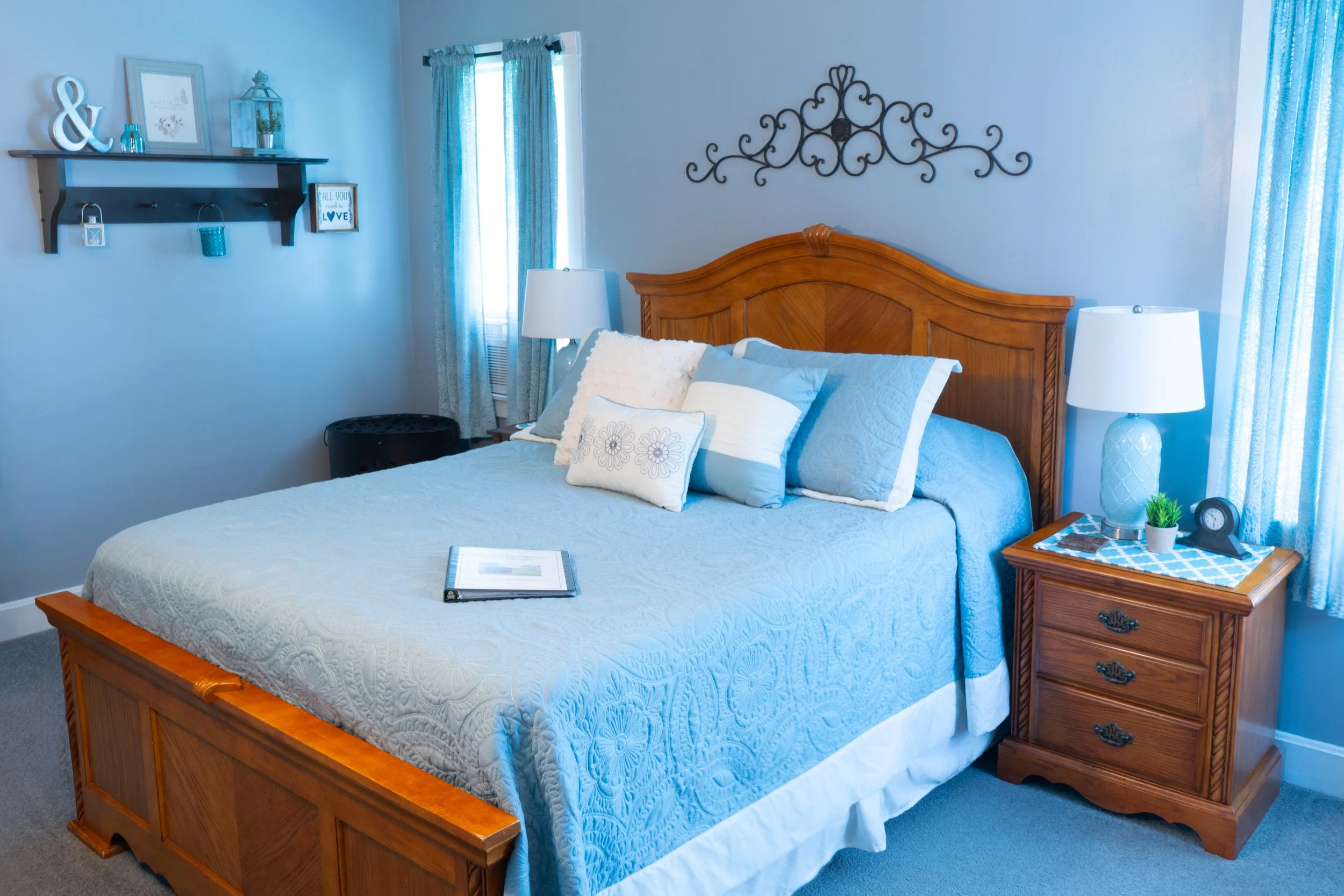 A bedroom with a wooden bed frame, blue quilt, and multiple pillows, flanked by two nightstands with lamps. Light blue curtains and decorative wall shelf with picture frames are visible.