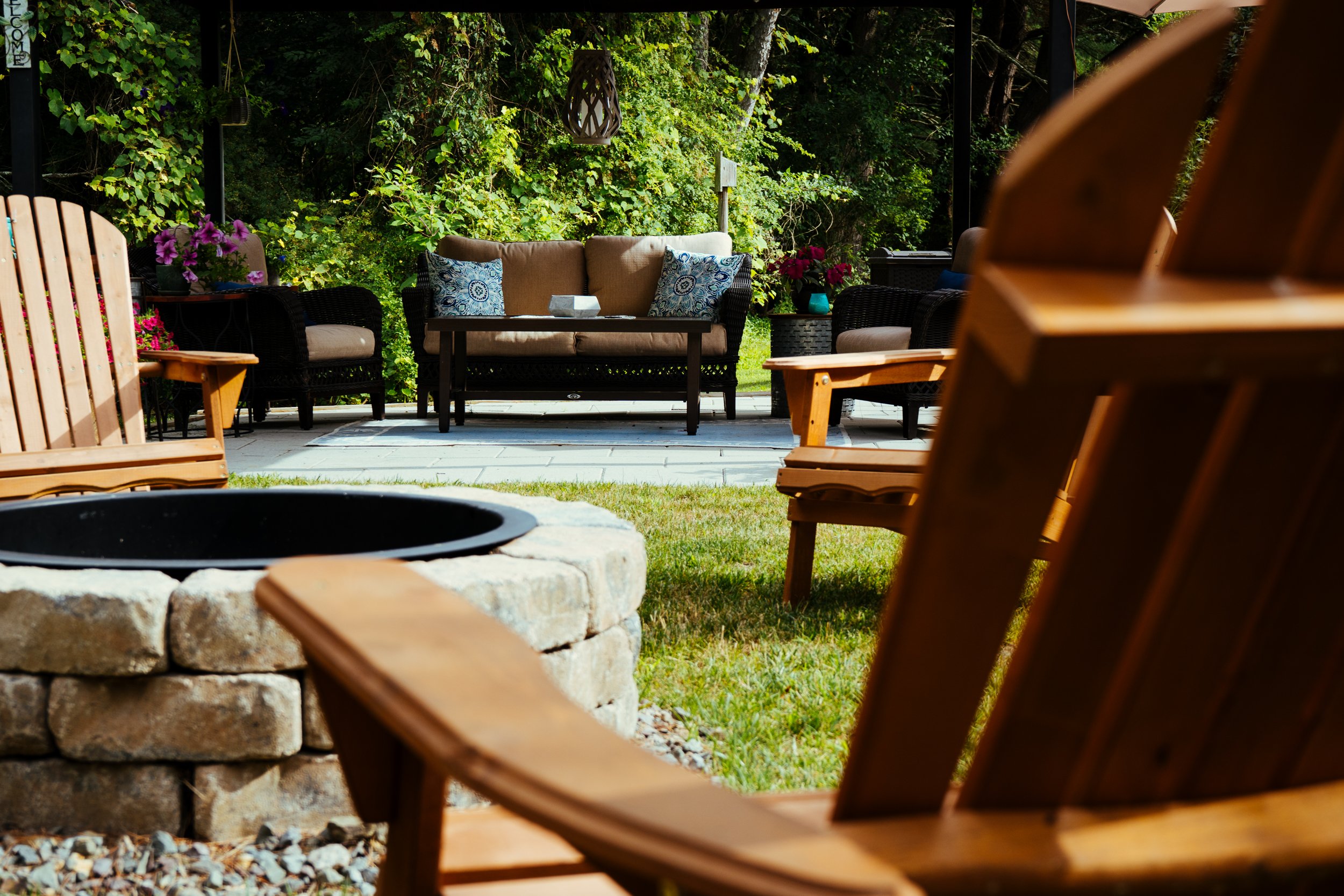 A backyard patio with wooden chairs, a fire pit, and a cushioned sofa set with pillows, surrounded by greenery.
