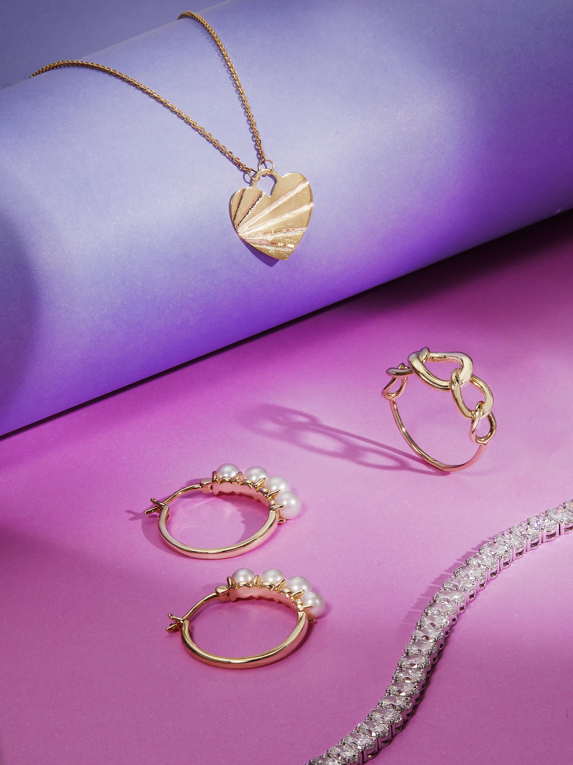 Gold jewelry set including a heart-shaped pendant necklace, pearly hoop earrings, a chain-link ring, and a rhinestone bracelet, displayed on colorful purple and pink backgrounds.