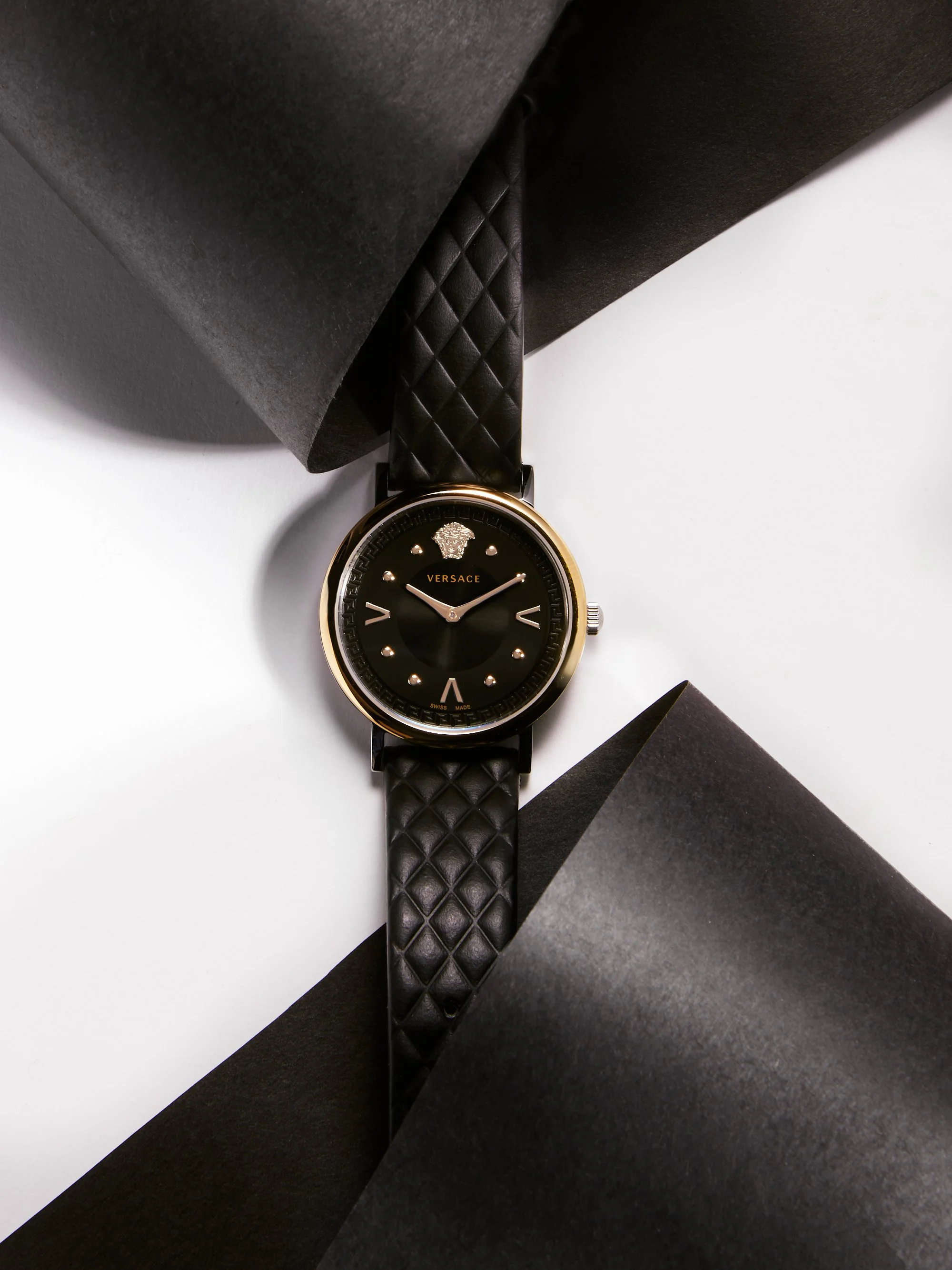 A black Versace watch with a gold bezel, black dial with gold markers, positioned on black paper against a white background.